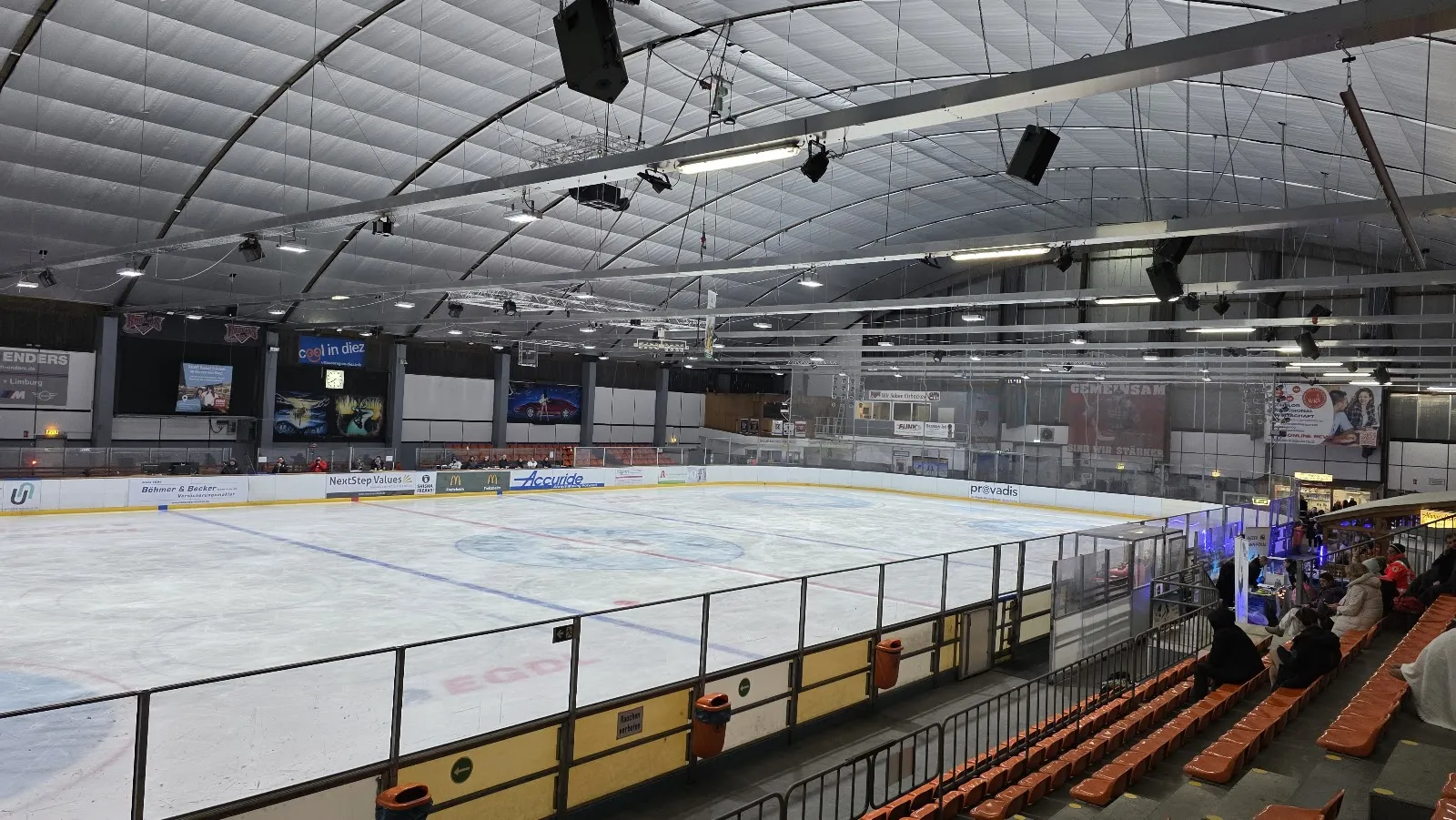 The ice sheet seen from a row of orange seats under the hall's curved, fabric-lined roof, with boards and sponsor panels along the far side. — Eissportzentrum Diez (Am Heckenweg)