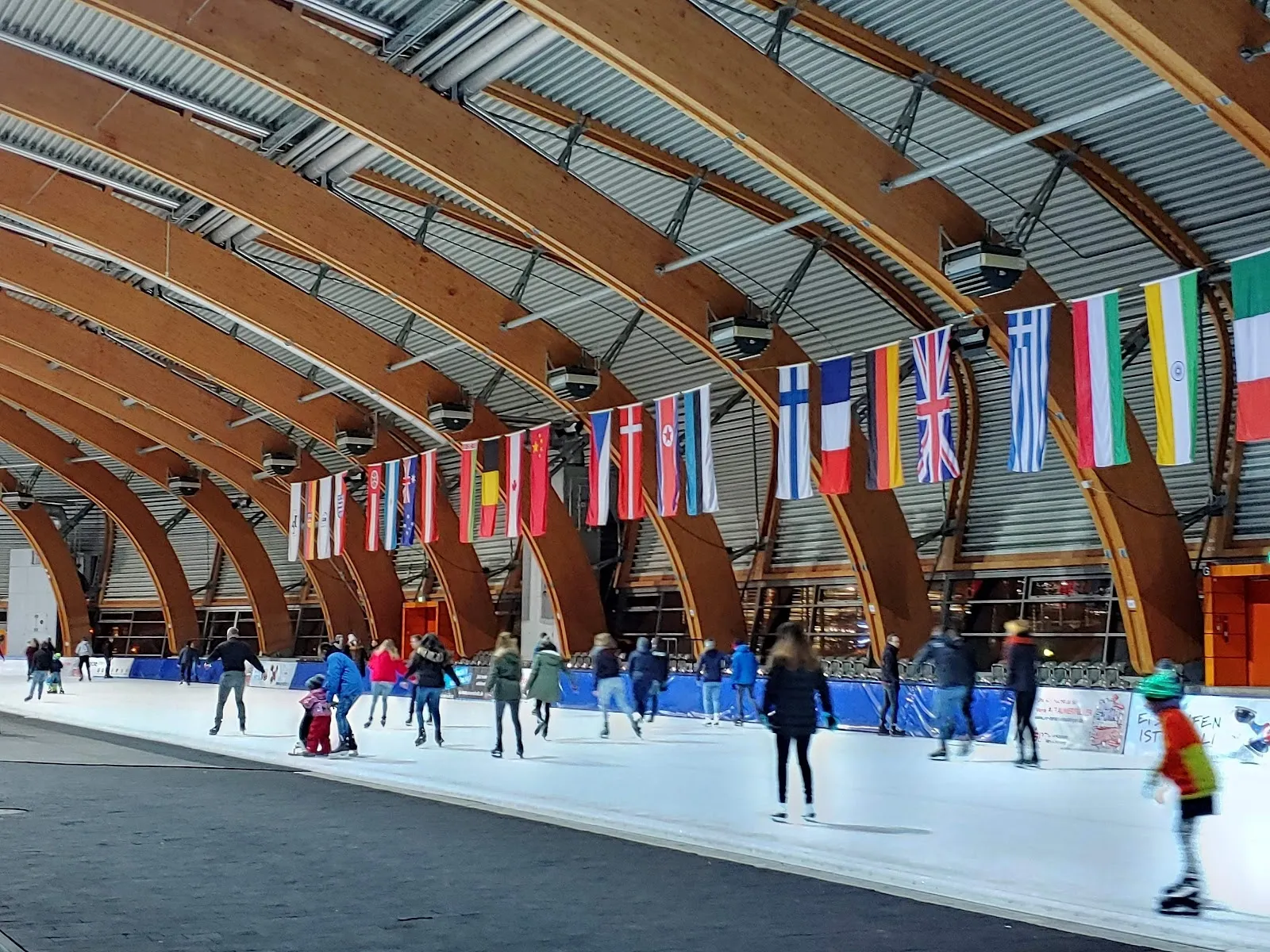 Publikumslauf unter den geschwungenen Holzbindern der Erfurter Eishalle, mit einer Reihe von Länderflaggen entlang der Bande. – Eissportzentrum Erfurt (Gunda-Niemann-Stirnemann-Halle)