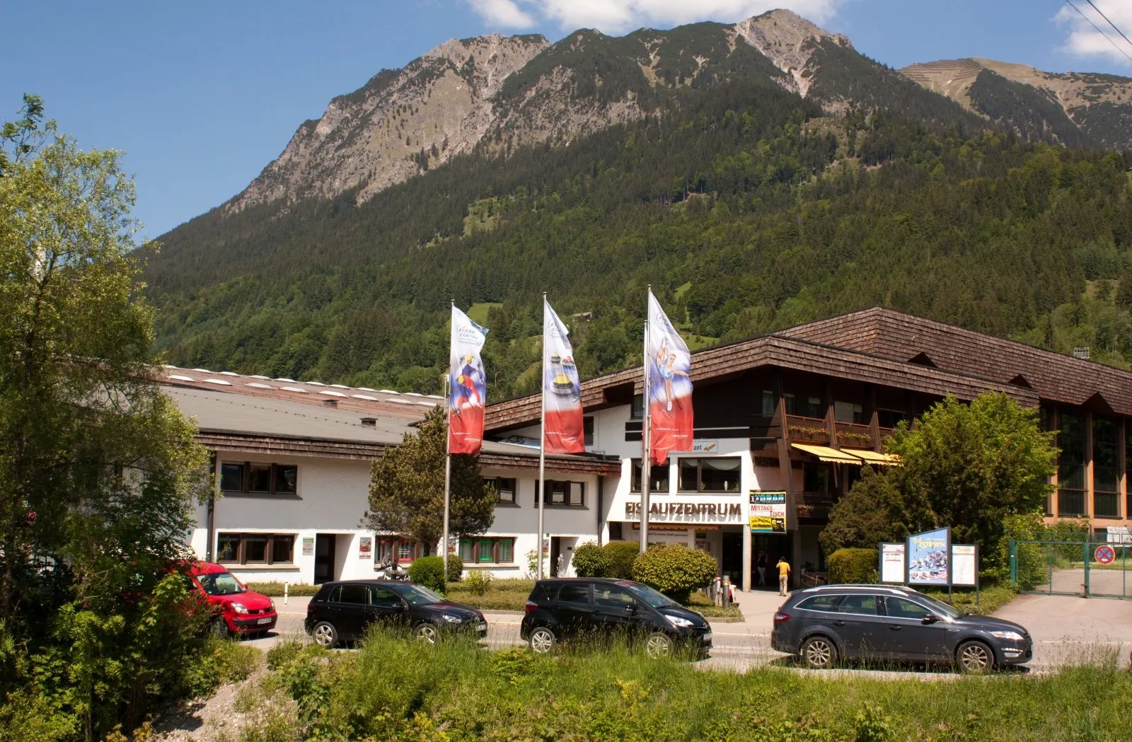 Die Vorderseite des Eissportzentrums Oberstdorf mit ihrer Fahnenreihe und geparkten Autos, dahinter die Allgäuer Berge. – Eissportzentrum Oberstdorf