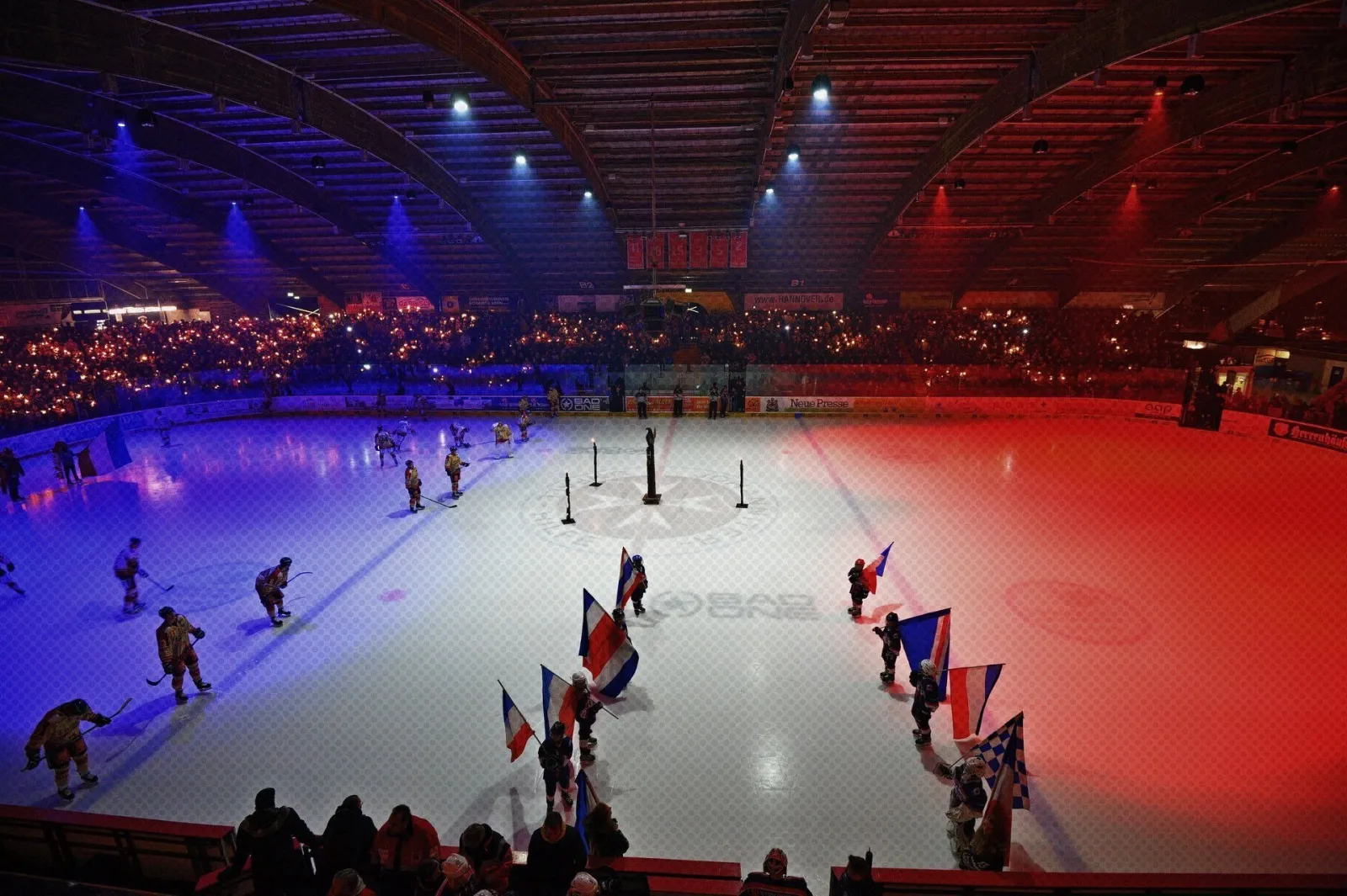 The Pferdeturm stadium lit in blue and red, skaters carrying flags across the ice with the stands behind. — Eisstadion am Pferdeturm