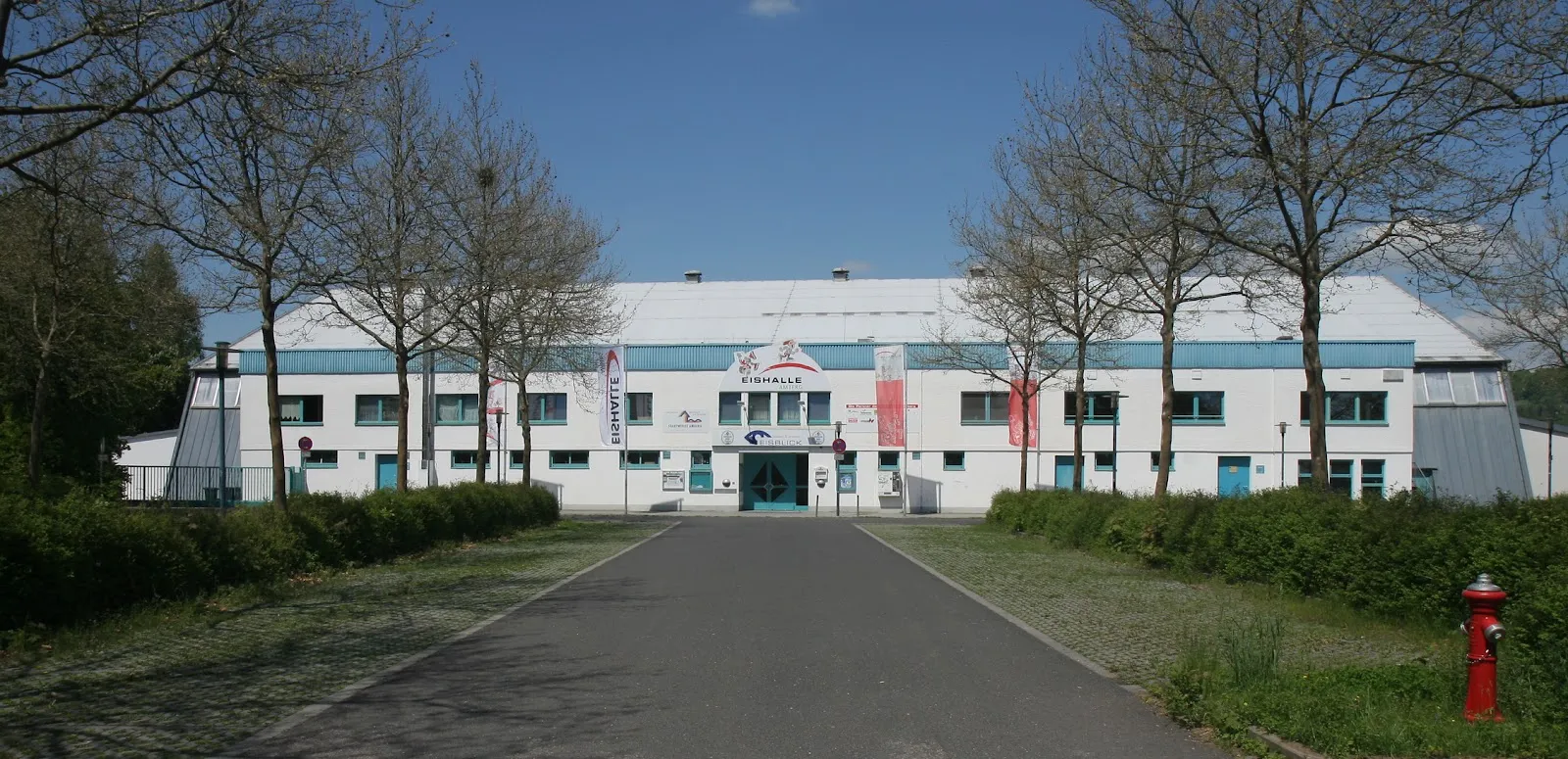 The long white ice hall with turquoise trim seen from the tree-lined approach road, its entrance and banners at the centre. — Eisstadion am Schanzl