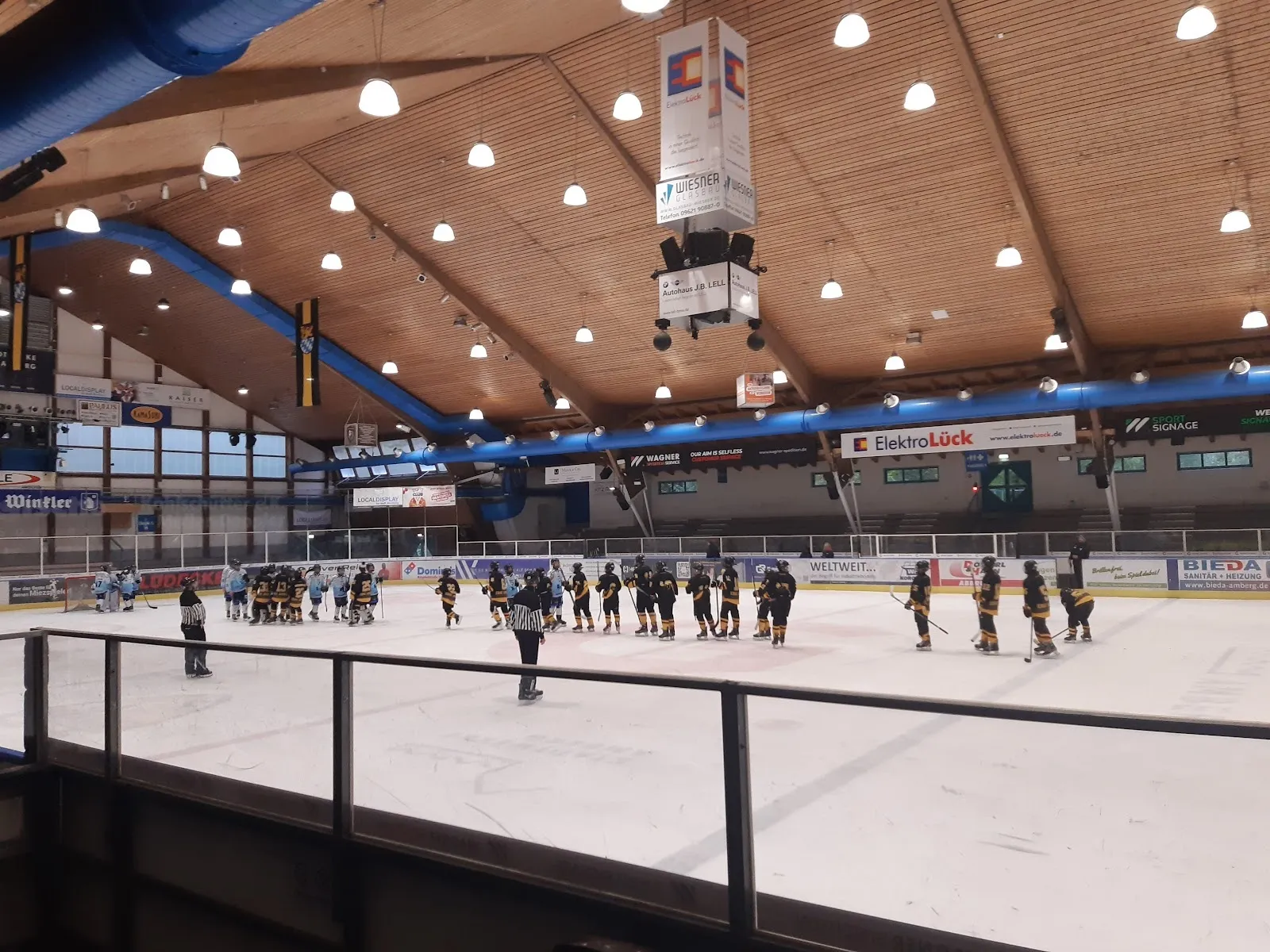 The ice sheet at the Amberg Eisstadion with a team lined up on the ice, seen over the rail under the timber roof and blue steel girders. — Eisstadion am Schanzl