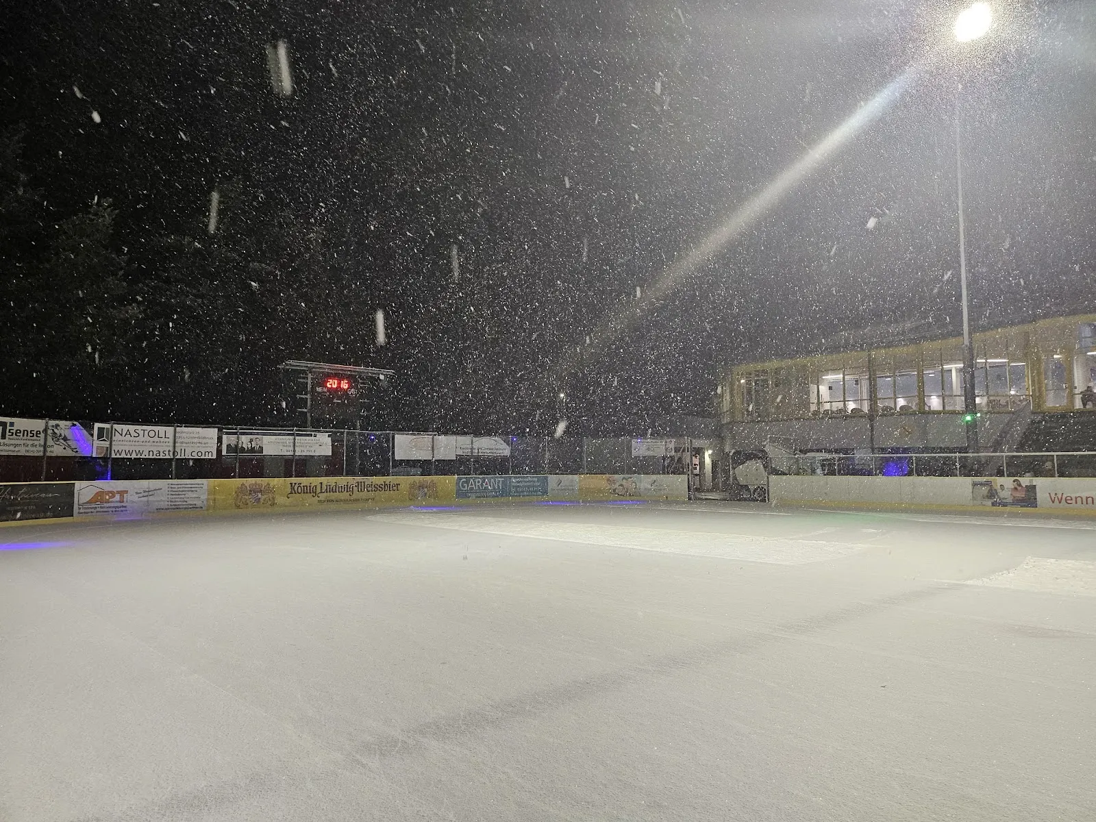 The open-air ice surface floodlit at night in falling snow, with sponsor boards along the far side and the lit stand building beyond. — Eisstadion der AmperOase Fürstenfeldbruck