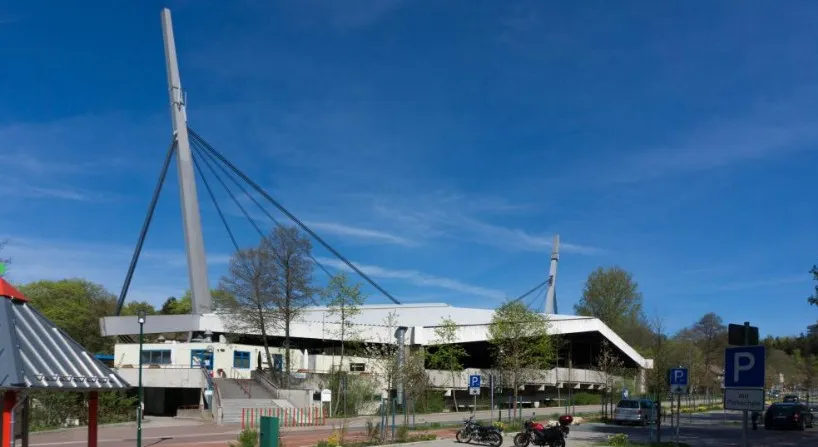 The stadium's white roof hung from two tall steel pylons with bundled cables, seen from the street beside the car park under a blue sky. — Eisstadion Braunlage
