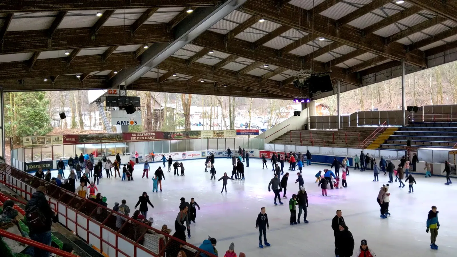 A public skating session in the covered stadium: skaters spread across the ice under the timber-and-steel roof, with the stands along the side. — Eisstadion Braunlage