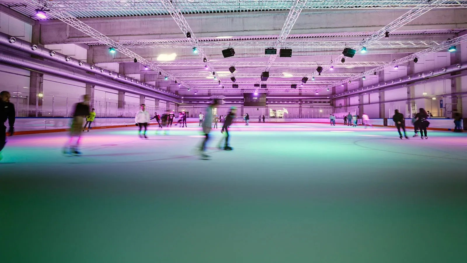 A public session in the Brehmstrasse hall, skaters crossing the ice under pink and green lighting with the open steel roof above. — Eisstadion Brehmstraße