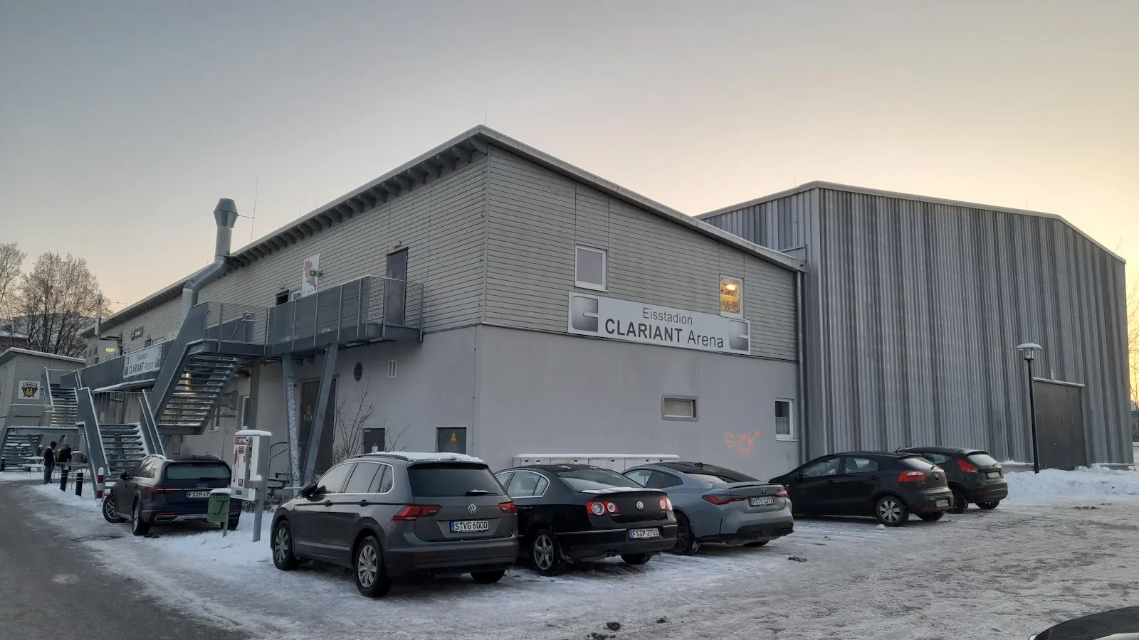 Die graue Seite des Eisstadions Clariant Arena in Moosburg mit dem Namensschild an der Wand und im Schnee davor parkenden Autos – Eisstadion Clariant Arena