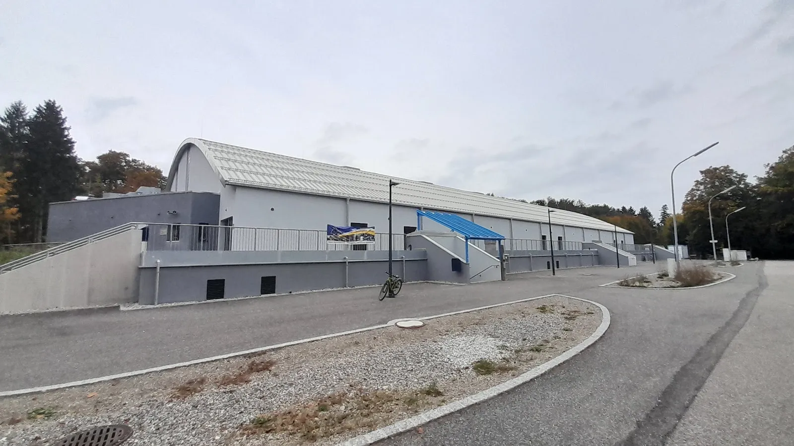 Die graue, verkleidete Halle mit gewölbtem Dach und blauem Eingangsvordach, gesehen von der geschwungenen Zufahrtsstraße unter bewölktem Himmel. – Heinz-Schneider-Eisstadion