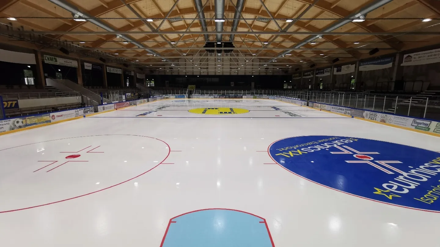 Die leere Eisfläche mit blauen und roten Markierungen und Sponsorenlogos, von einem Ende der Halle aus gesehen, unter einem Dach mit Holzbalken. – Heinz-Schneider-Eisstadion