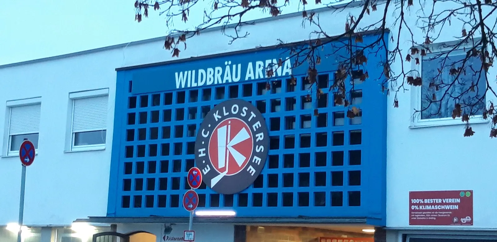 Die weiße Vorderwand der Arena mit einer blauen Gitterplatte mit dem Schriftzug Wildbräu Arena über dem Emblem des EHC Klostersee. – Eisstadion Grafing (Wildbräu Arena)