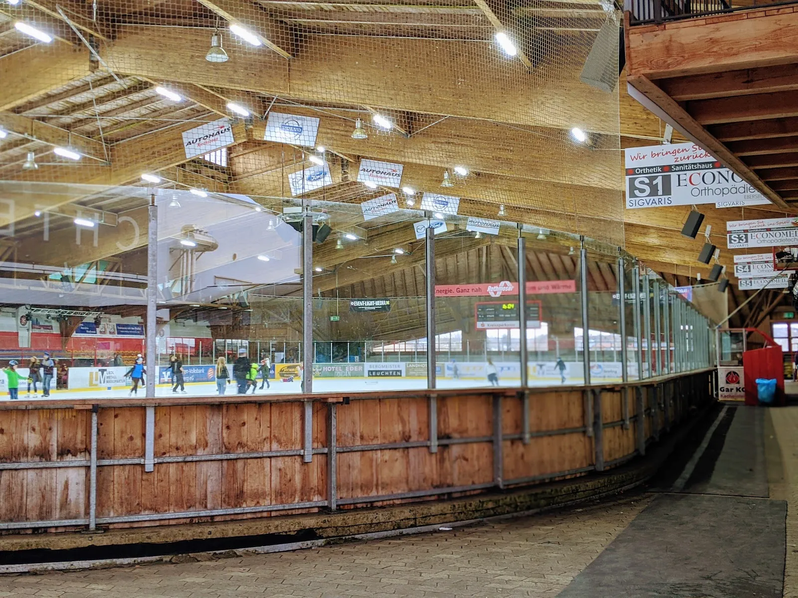 Die Eisfläche, hinter der geschwungenen Holzbande und den Glasscheiben gesehen, mit Läufern auf dem Eis und dem Holzdach der Arena, an dem Sponsorenbanner hängen. – Eisstadion Grafing (Wildbräu Arena)