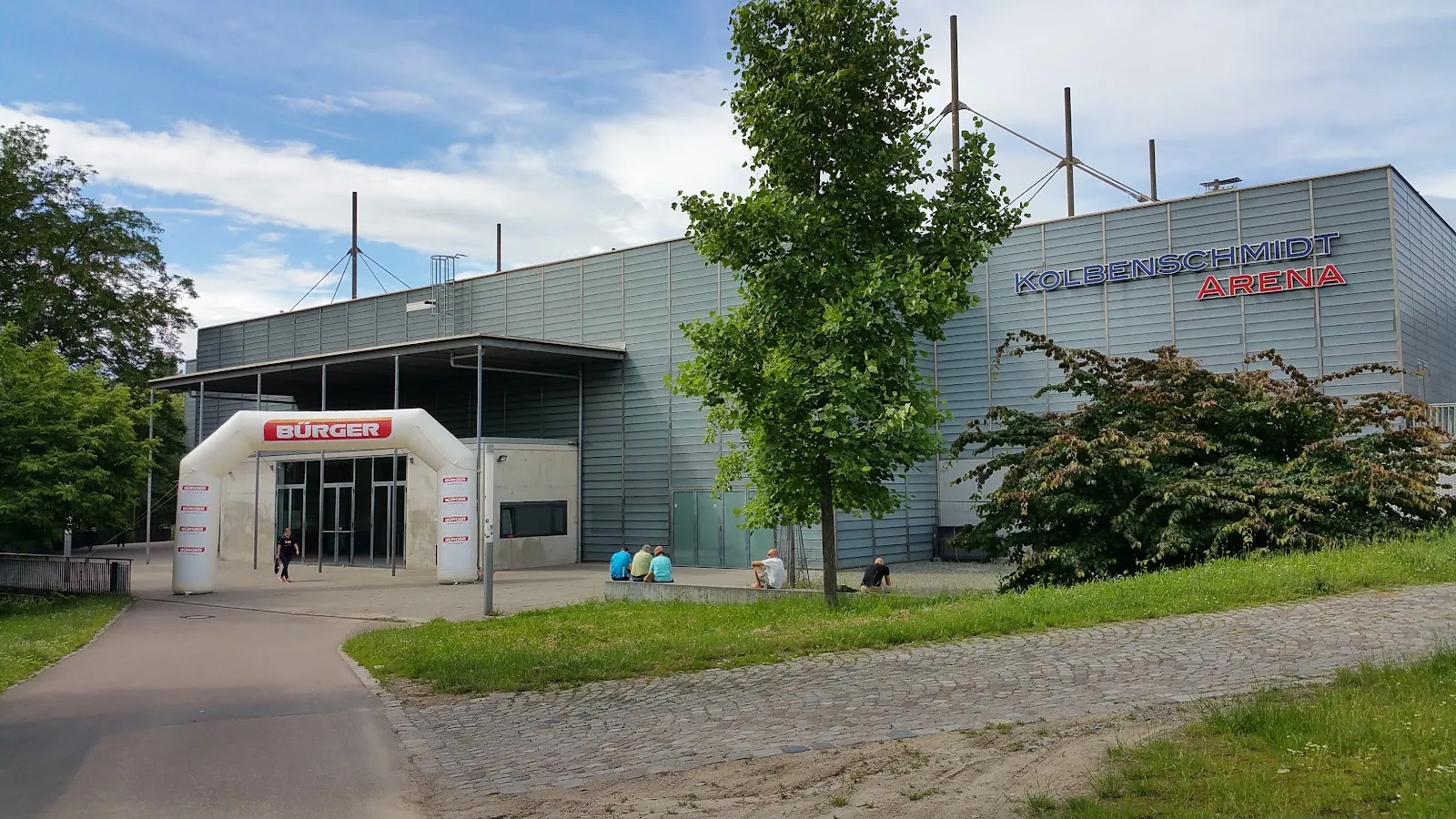 The arena's grey metal-clad front with its entrance and the Kolbenschmidt Arena lettering, set among trees and lawn. — Eisstadion Heilbronn