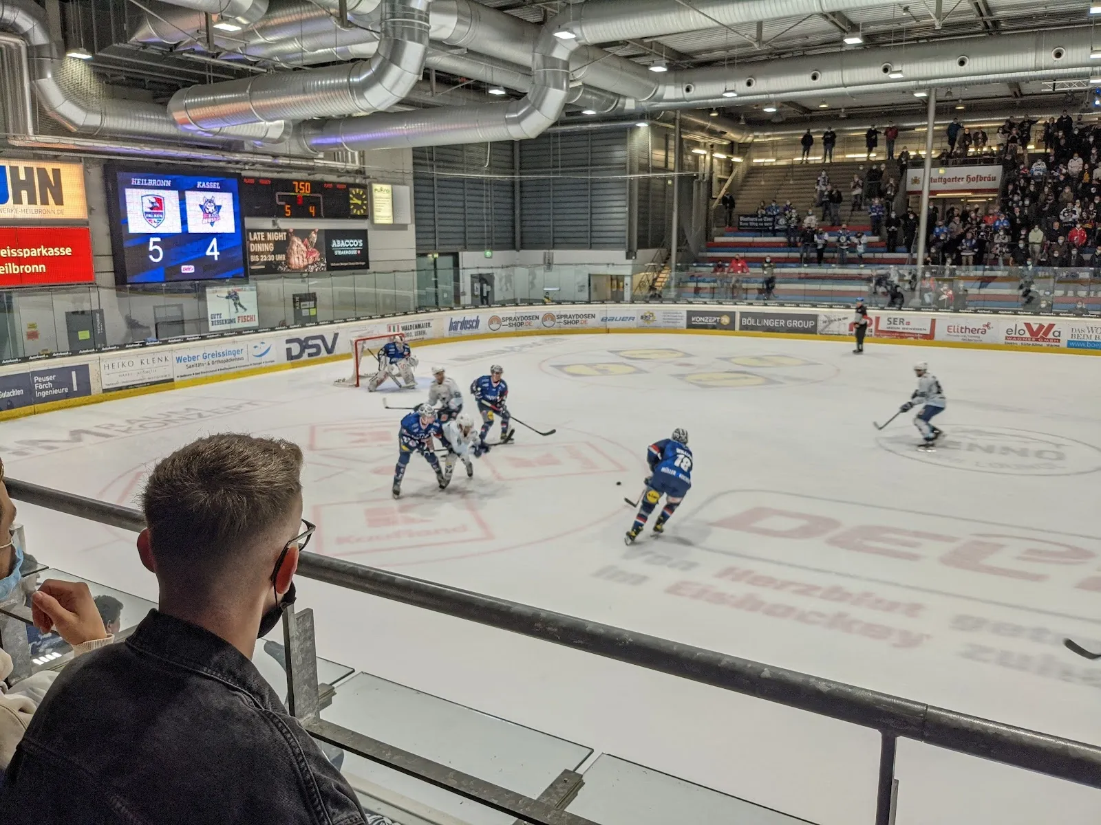 An ice hockey game in play at the Knorr Arena in Heilbronn, seen from the stands: the ice with players at a face-off, the scoreboard and the packed stands behind the boards. — Eisstadion Heilbronn