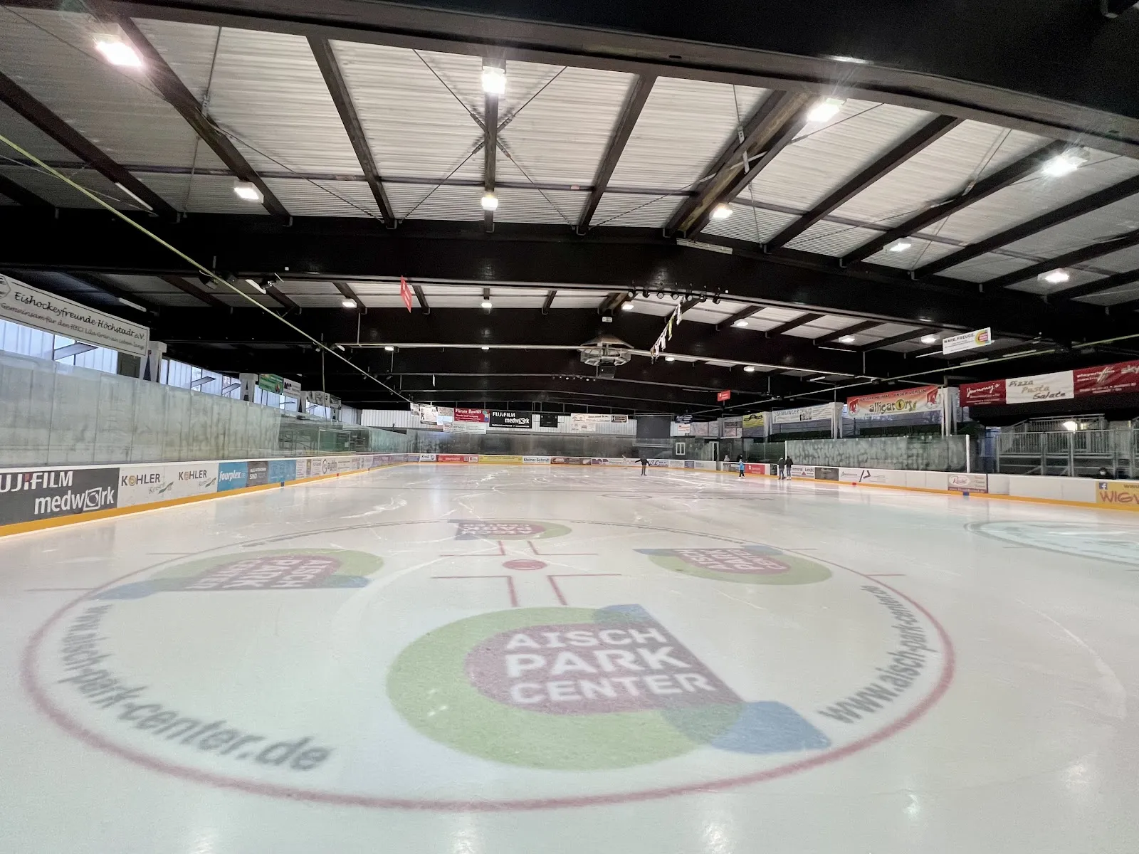 The empty ice sheet with the large centre-ice logo ringed in red, steel roof trusses overhead and the boards running into the distance. — Eisstadion Höchstadt an der Aisch