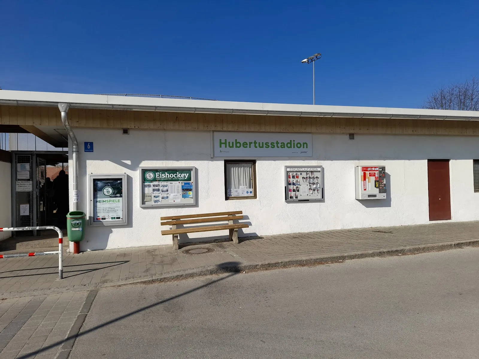 Das niedrige weiße Vereinsgebäude mit dem Schild Hubertusstadion, Aushängen und einer Bank an der Straße unter klarem blauem Himmel. – Eisstadion Holzkirchen (Hubertus-Stadion)