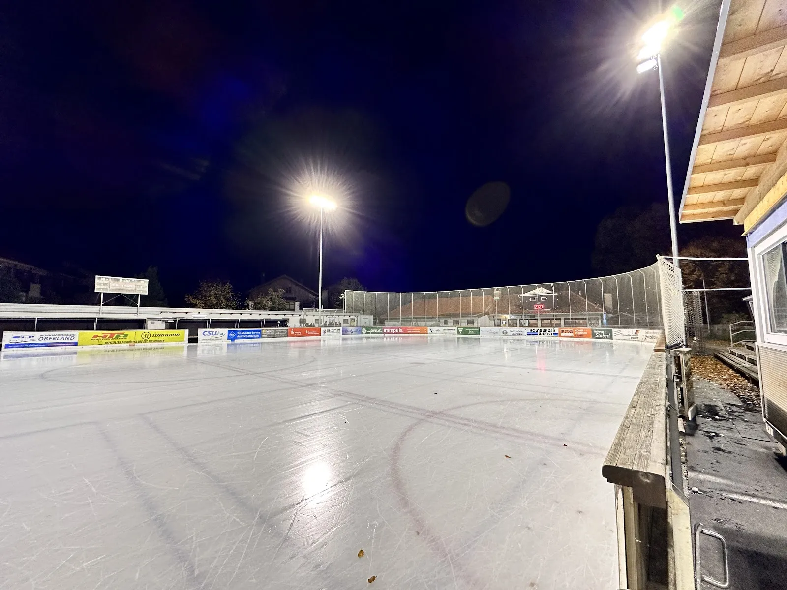 Die offene Eisfläche im Flutlicht vor dunkelblauem Himmel, im Vordergrund vom Schlittschuhlaufen zerfurchtes Eis, an der gegenüberliegenden Seite Werbebanden und Netze. – Eisstadion Holzkirchen (Hubertus-Stadion)
