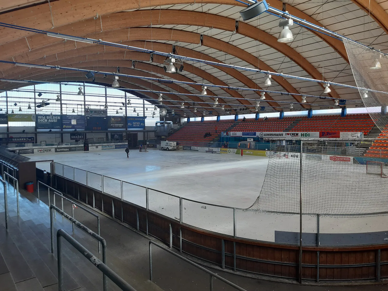 Die Eisfläche, von den Rängen aus gesehen, unter dem weit geschwungenen Brettschichtholz-Bogendach der Arena, mit der gegenüberliegenden Tribüne und einer verglasten Giebelwand hinter den Banden. – Eisstadion Kempten