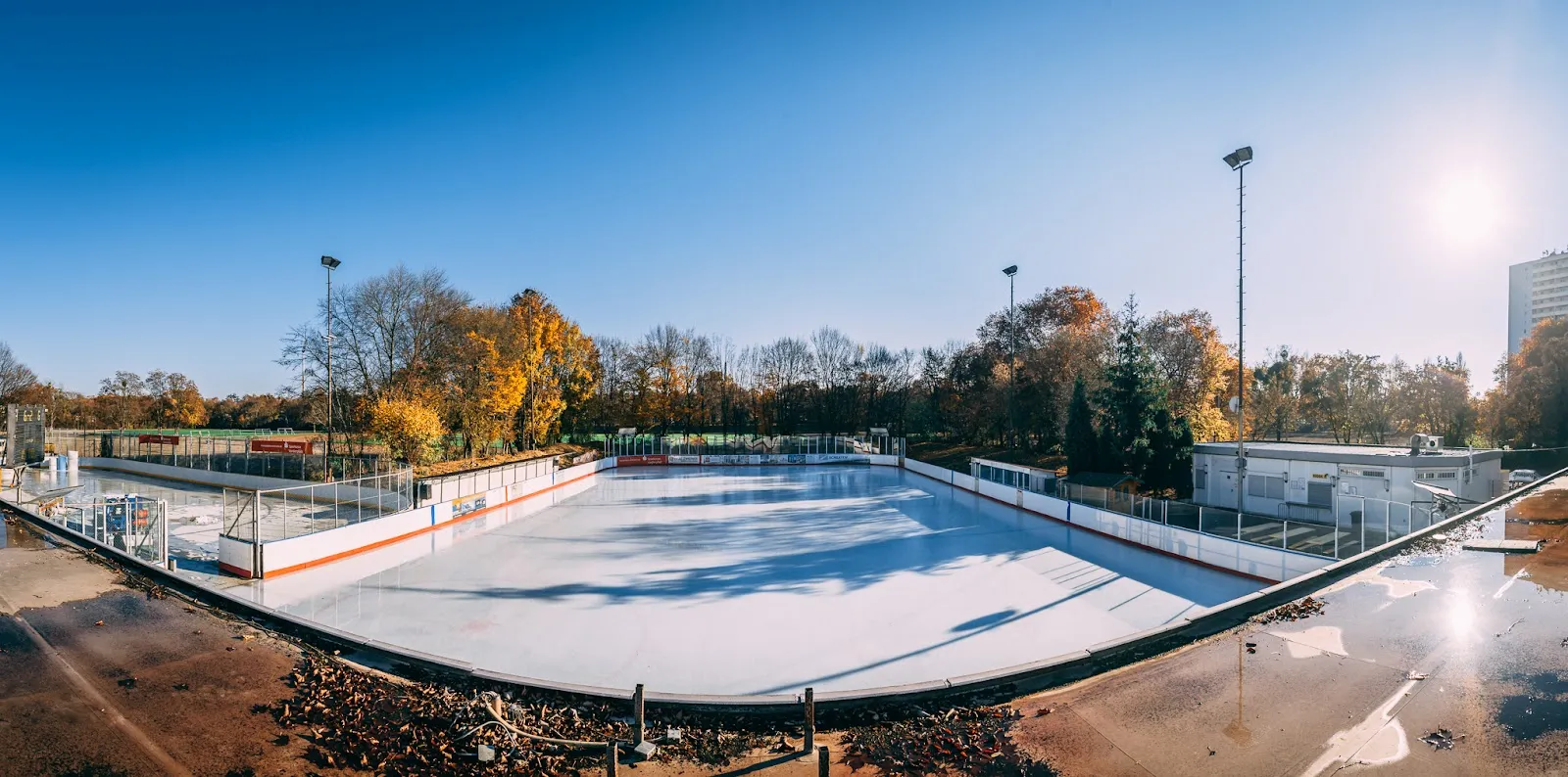 Ein Panoramablick über die Freiluft-Eisbahn in der Wintersonne, mit Banden, Flutlichtmasten und den herbstlichen Bäumen des umgebenden Parks. – Eisstadion Ludwigshafen (ERCL)