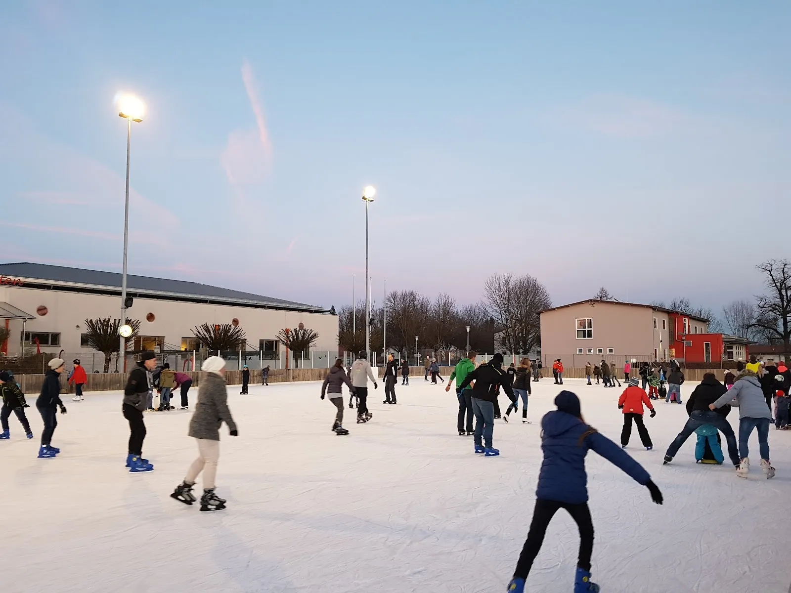 The open-air Eisstadion in Mühldorf at dusk with skaters across the ice, the floodlights and the low buildings along the far side. — Eisstadion Mühldorf (Kunsteisbahn)