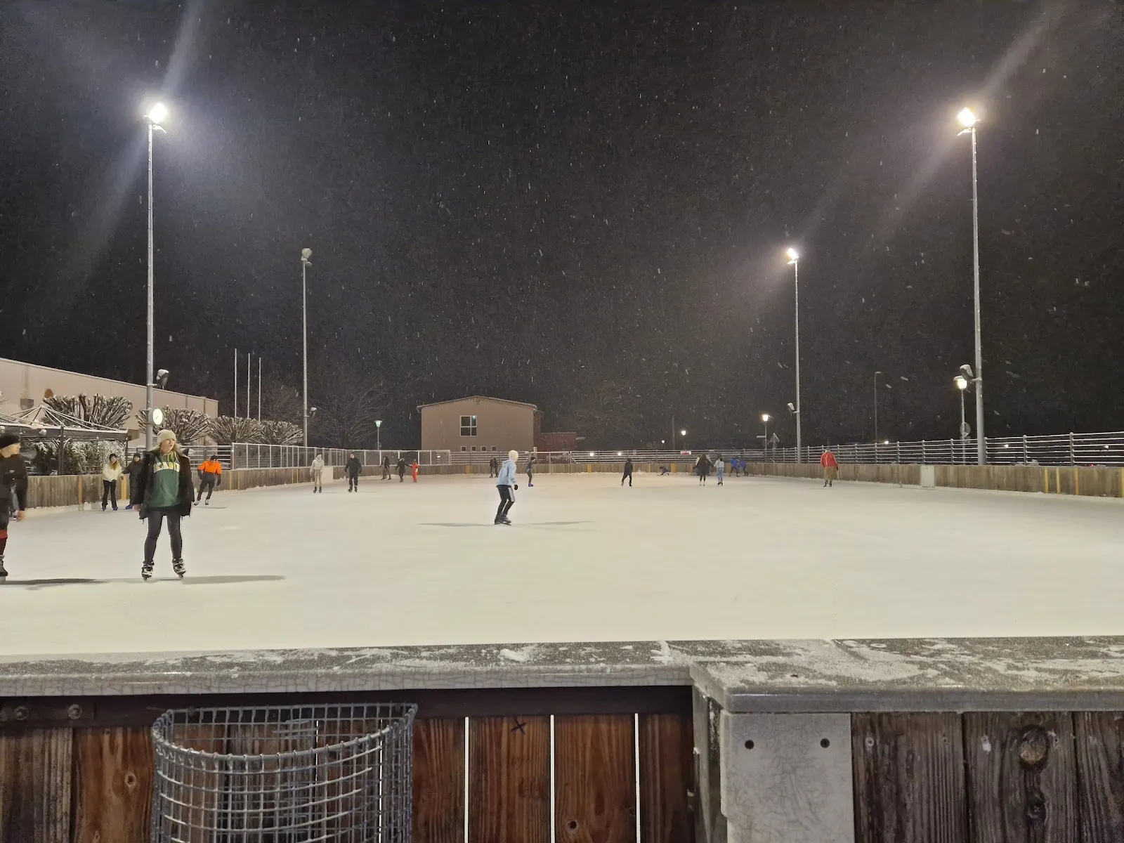 The large open-air rink floodlit at night in light snowfall, with a few skaters and the wooden boards in the foreground and a low building at the far side. — Eisstadion Mühldorf (Kunsteisbahn)