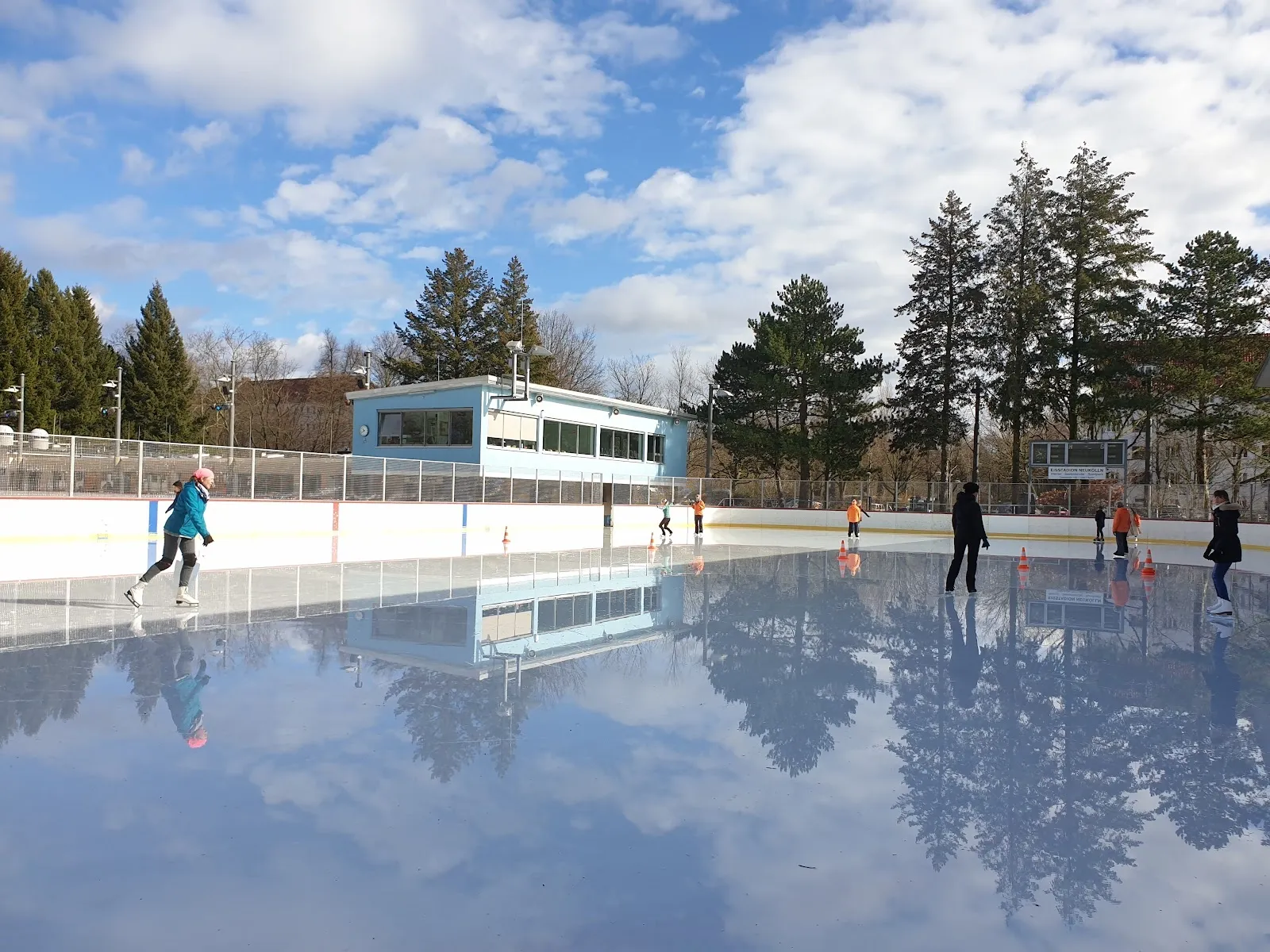 The open-air Eisstadion Neukölln in Berlin on a winter day, with skaters on the ice and the low rink-side building and trees behind. — Eisstadion Neukölln