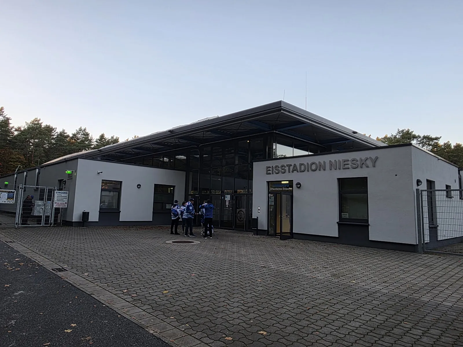 The low entrance building with a projecting roof and the Eisstadion Niesky lettering on its white wall, seen across a paved forecourt at dusk. — Eisstadion Niesky