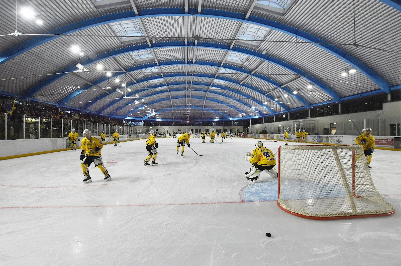 A hockey game in play on the ice of the Niesky ice stadium, seen from the boards under the blue arched roof. — Eisstadion Niesky