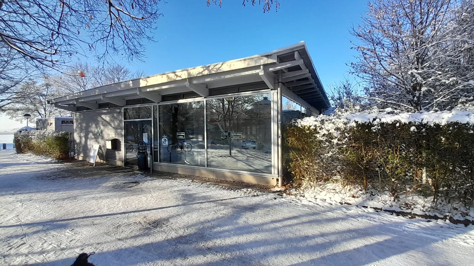 Der niedrige verglaste Eingangspavillon mit einem flachen, vorkragenden Dach, Schnee auf dem Boden und auf der Hecke daneben, unter klarem blauem Himmel. – Eisstadion Ottobrunn