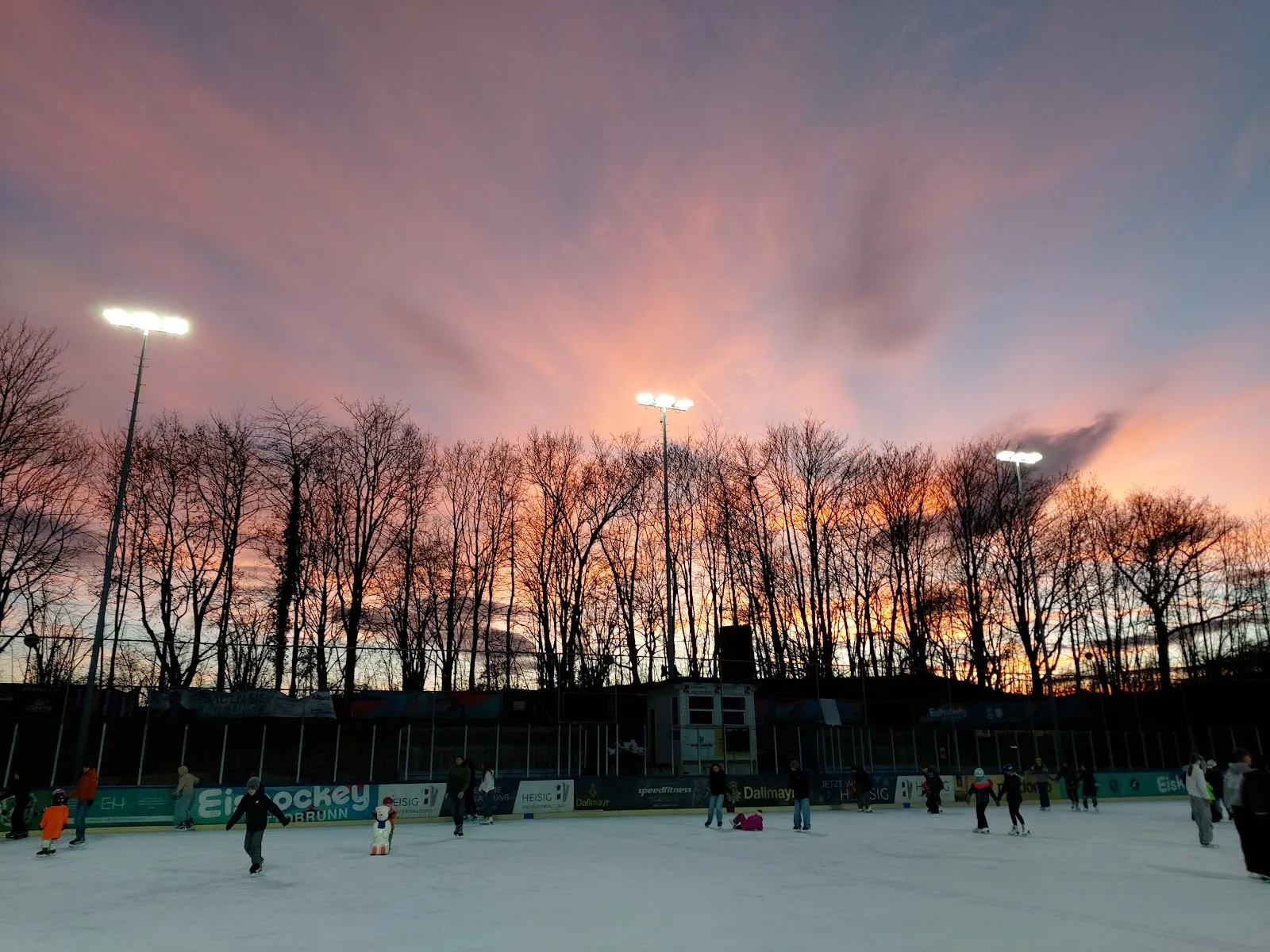 Das Freiluft-Eis bei Sonnenuntergang, mit einigen Läufern an den hinteren Banden, Flutlichtmasten und einer Reihe kahler Bäume als Silhouetten vor einem rosa-orangen Himmel. – Eisstadion Ottobrunn