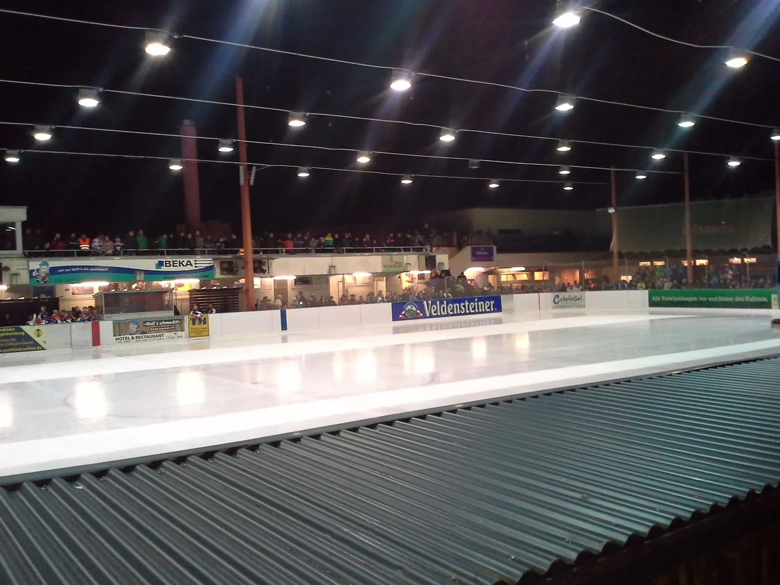 Die Bahn bei Nacht im Licht der Halle, mit weißem Eis, den Banden und den blauen Werbetafeln; keine Personen auf dem Eis. – Eisstadion Pegnitz