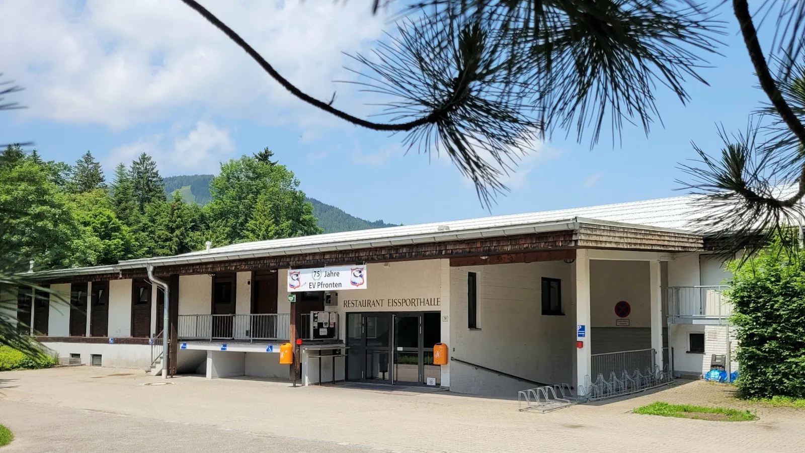 Das niedrige Eingangsgebäude des Eisstadions mit dem Schild des Restaurants und einem überdachten Gang, umgeben von Kiefern und einem Kiesvorplatz. – Eisstadion Pfronten