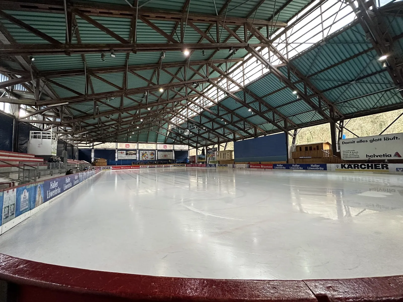 The empty ice sheet of the Eisstadion Polarion seen from the boards, under the open steel-trussed roof with its green translucent panels — Eisstadion Polarion