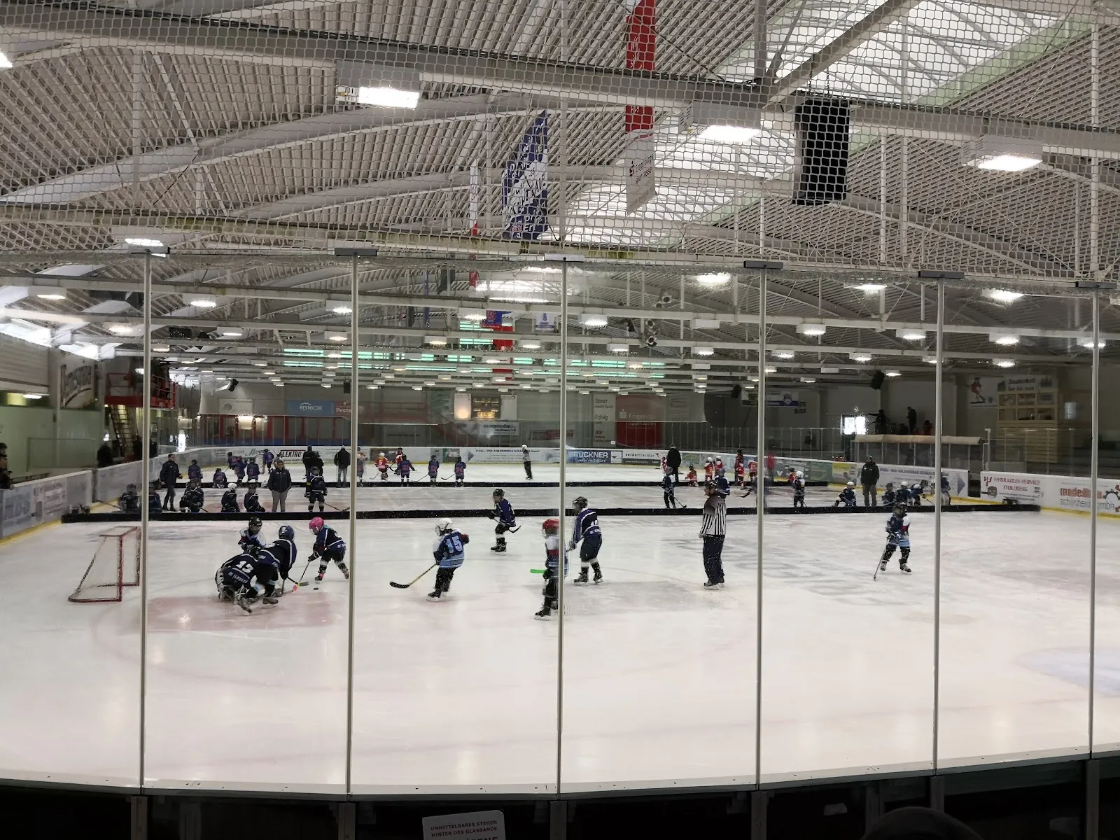 Die gesamte Eisfläche des Eisstadions Schönheide, von den Rängen durch die Scheiben gesehen, unter einem weißen Stahlfachwerkdach, mit einer kleinen Nachwuchs-Eishockeygruppe weit hinten auf dem Eis (Name des Maps-Eintrags: Wolfsbau Schönheide) – Eisstadion Schönheide