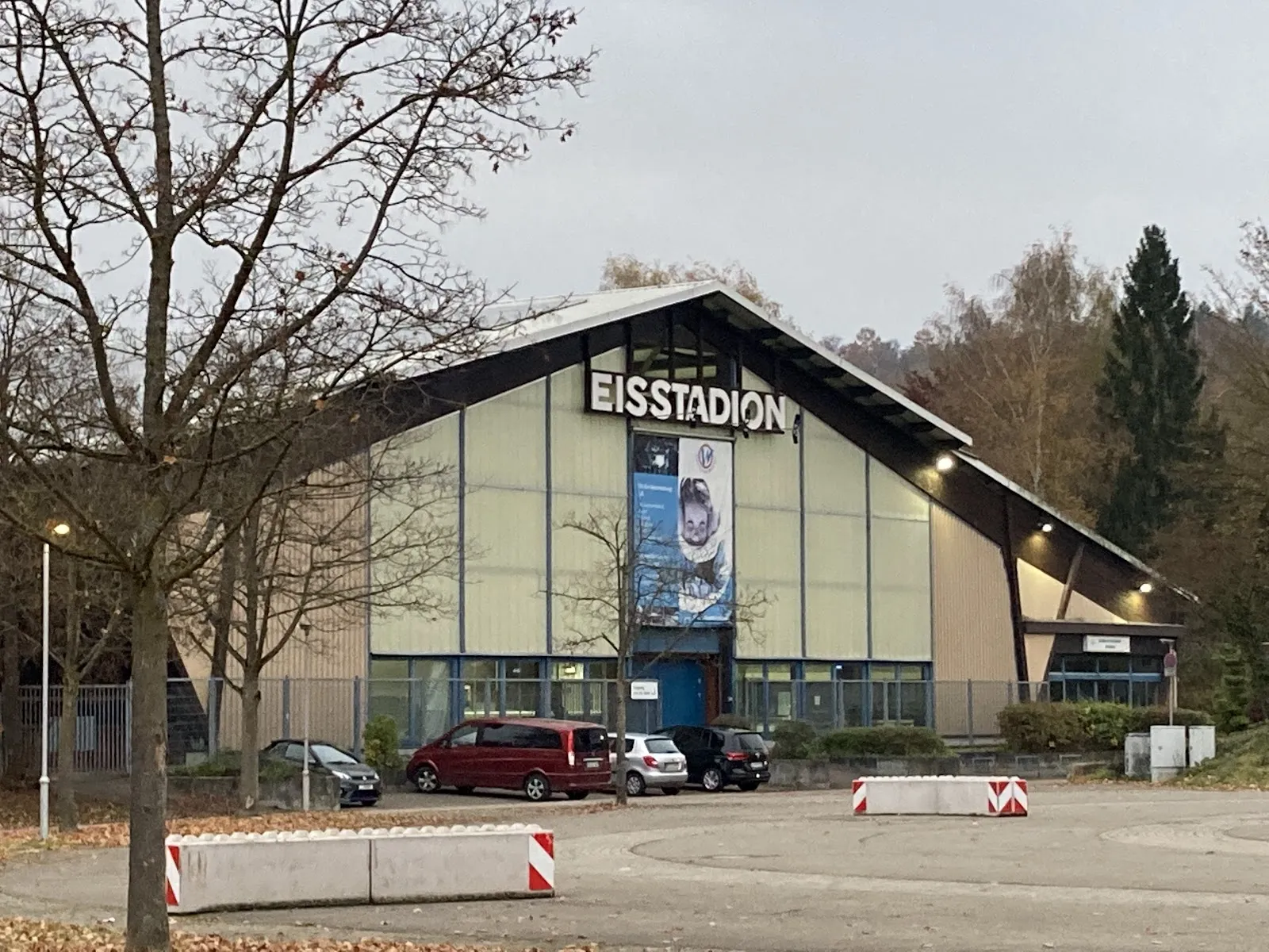 Die Giebelfront des Eisstadions Wernau mit dem Namen in großen Buchstaben über dem verglasten Eingang, über den Parkplatz hinweg gesehen – Eisstadion Wernau