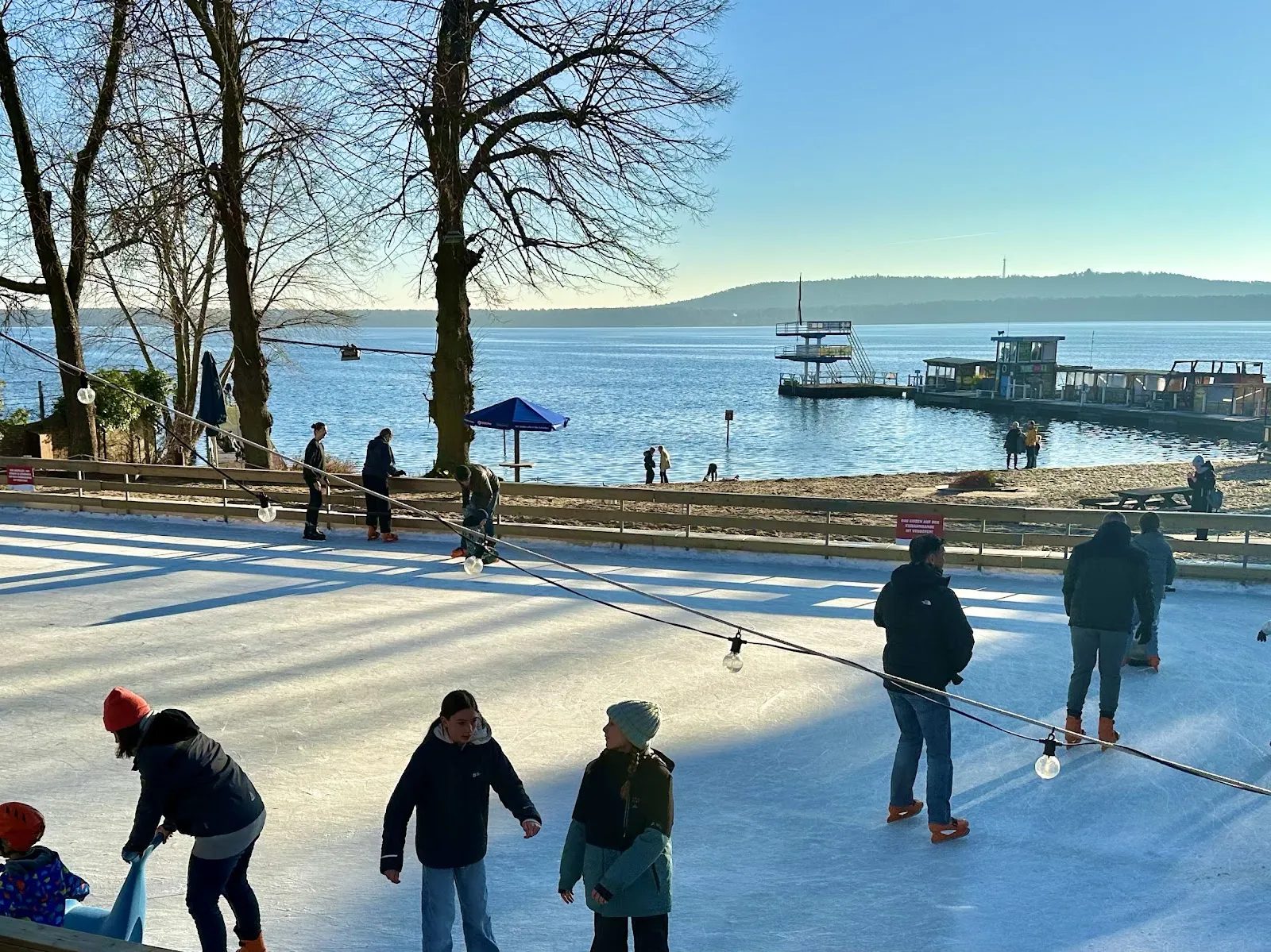 Die Eisstrand-Bahn im Strandbad Friedrichshagen am Ufer des Müggelsees, mit Schlittschuhläufern auf dem Eis in der Wintersonne. – Eisstrand Friedrichshagen