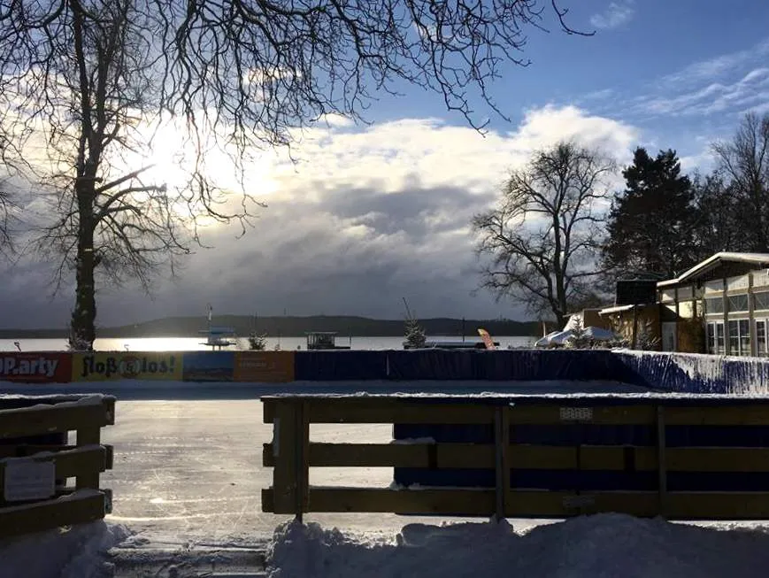 Die Holzbanden und blauen Werbetafeln der Eisbahn am See in der Wintersonne, dahinter das Müggelsee-Ufer und ein Kiosk. – Eisstrand Friedrichshagen