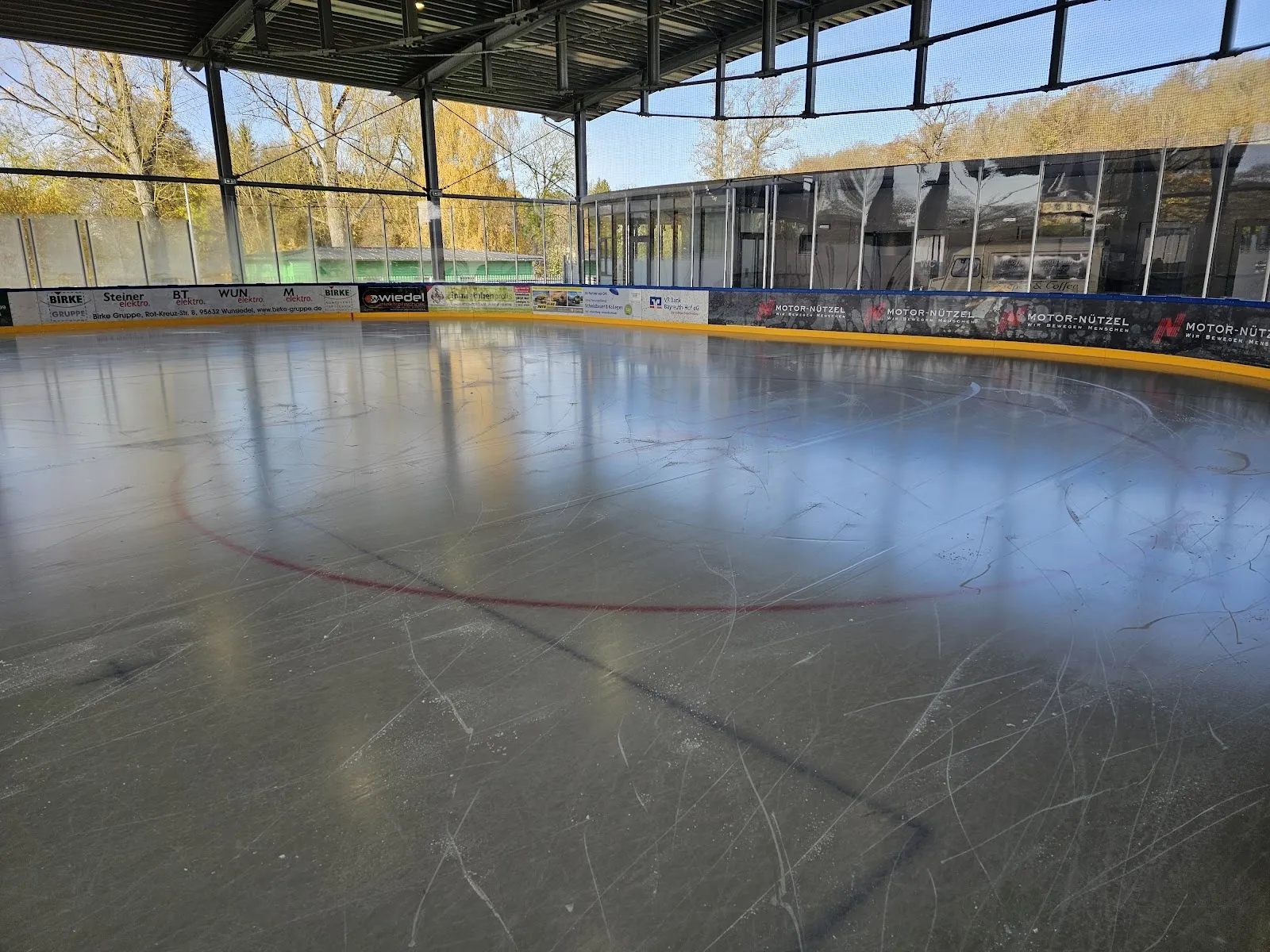 Die leere Eisfläche des Eisteichs in Hof, einer überdachten, seitlich offenen Bahn, mit der Bande und dem Dachrand. – Eisteich Hof