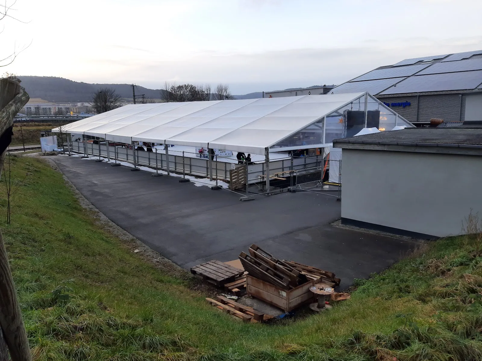 Die lange weiße Zelthalle mit Glasfronten, die die Eisbahn beherbergt, neben einer Grasböschung, mit Holzhütten am Ende und den Hügeln des Saaletals dahinter. – Eiswelt Jena