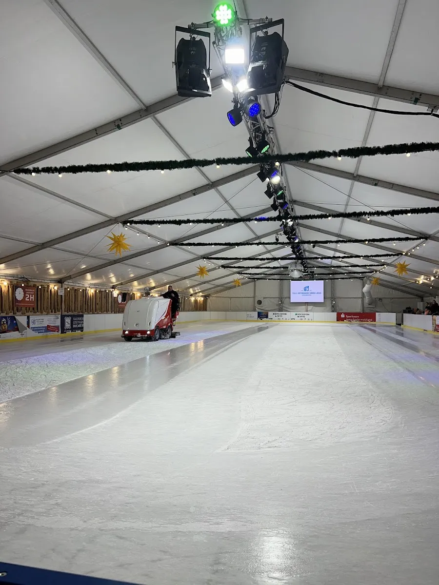 Die lange, leere Eisfläche im Zelt, am hinteren Ende frisch von einer roten Eisaufbereitungsmaschine bearbeitet, unter einem weißen Dach mit Girlanden und Scheinwerfern. – Eiswelt Jena