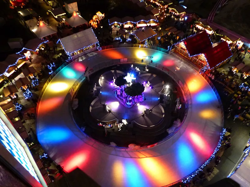 Der farbig beleuchtete Ring um den Neptunbrunnen bei Nacht von oben, umgeben von den Buden des Weihnachtsmarkts. – Eiswelt am Neptunbrunnen