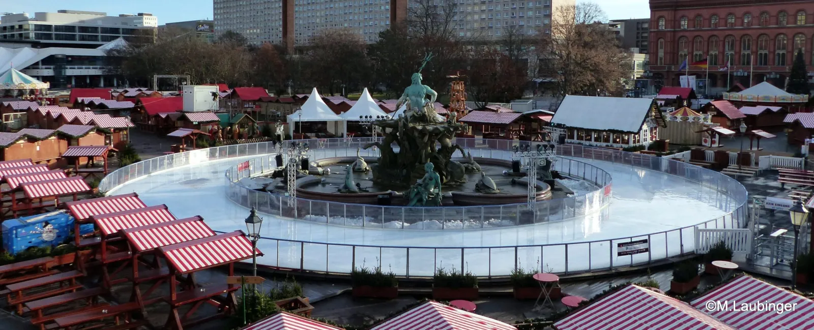 Die runde Eisbahn um den Neptunbrunnen mit einigen Schlittschuhläufern in der Ferne, umgeben von den rot-weißen Marktbuden und der Umgebung des Roten Rathauses. – Eiswelt am Neptunbrunnen