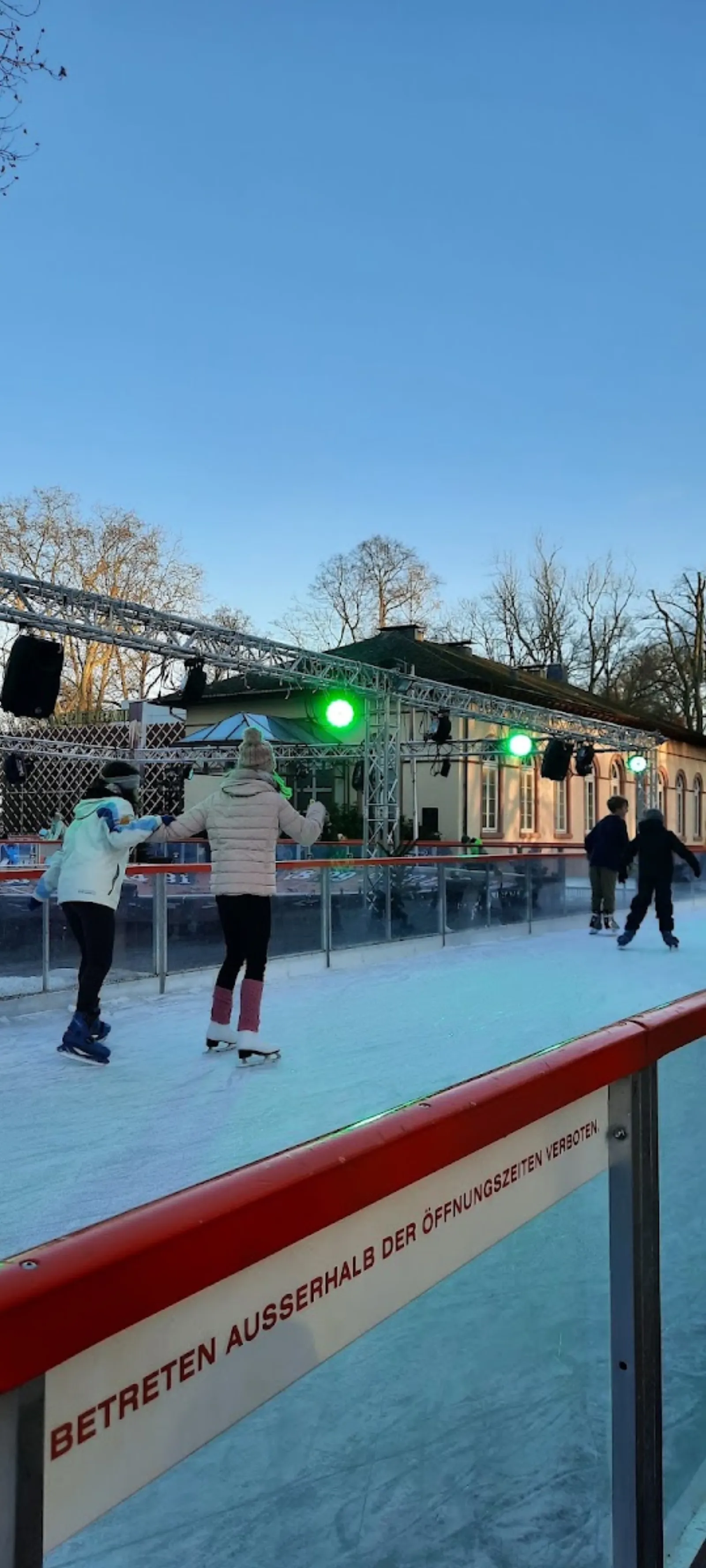Die Eiswinter-Bahn im Bad Homburger Kurpark, mit dem Pavillon und dem Lichtgerüst hinter den von hinten zu sehenden Schlittschuhläufern. – Bad Homburger Eiswinter