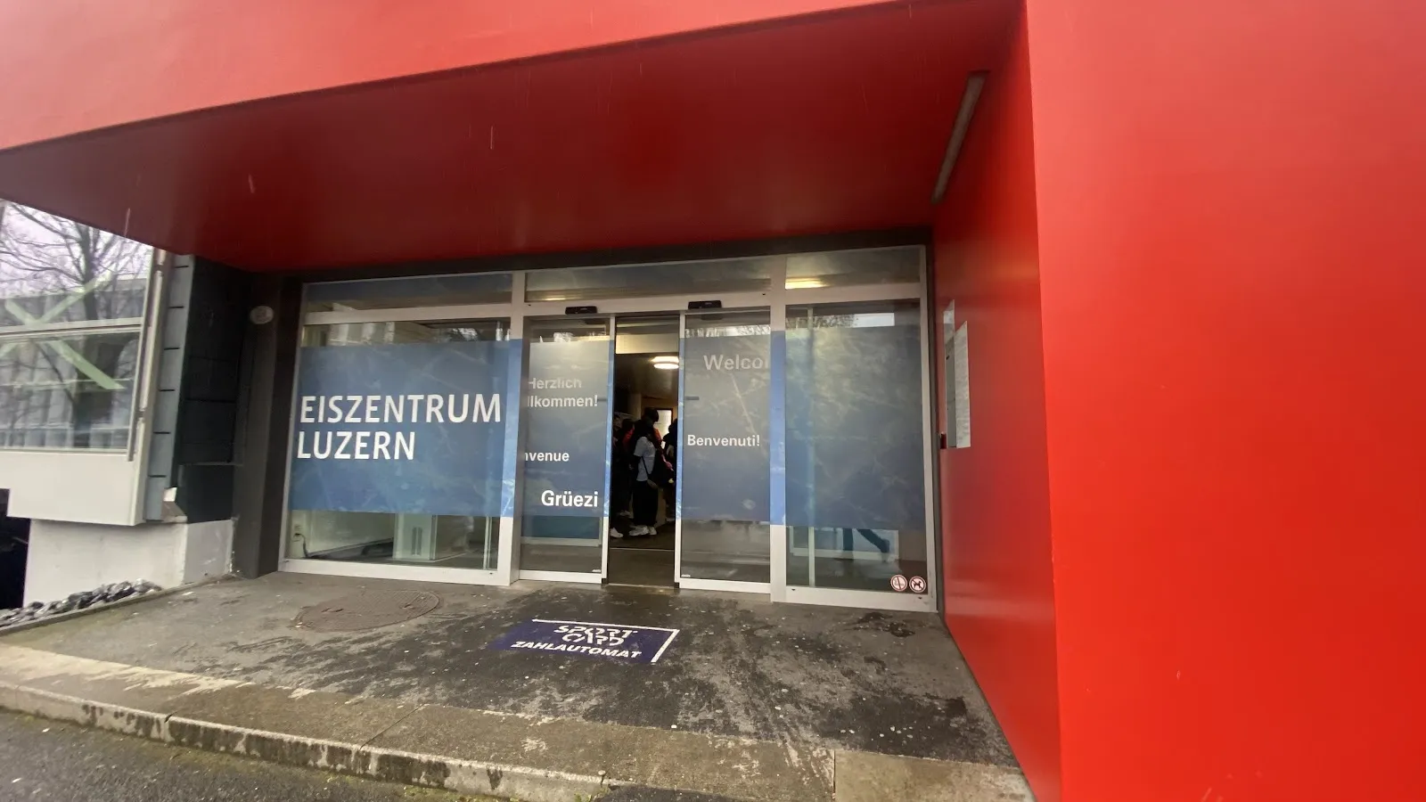 Die rote Front des Eiszentrums Luzern mit den gläsernen Eingangstüren und der abgeschrägten roten Fassade. – Eiszauber Luzern