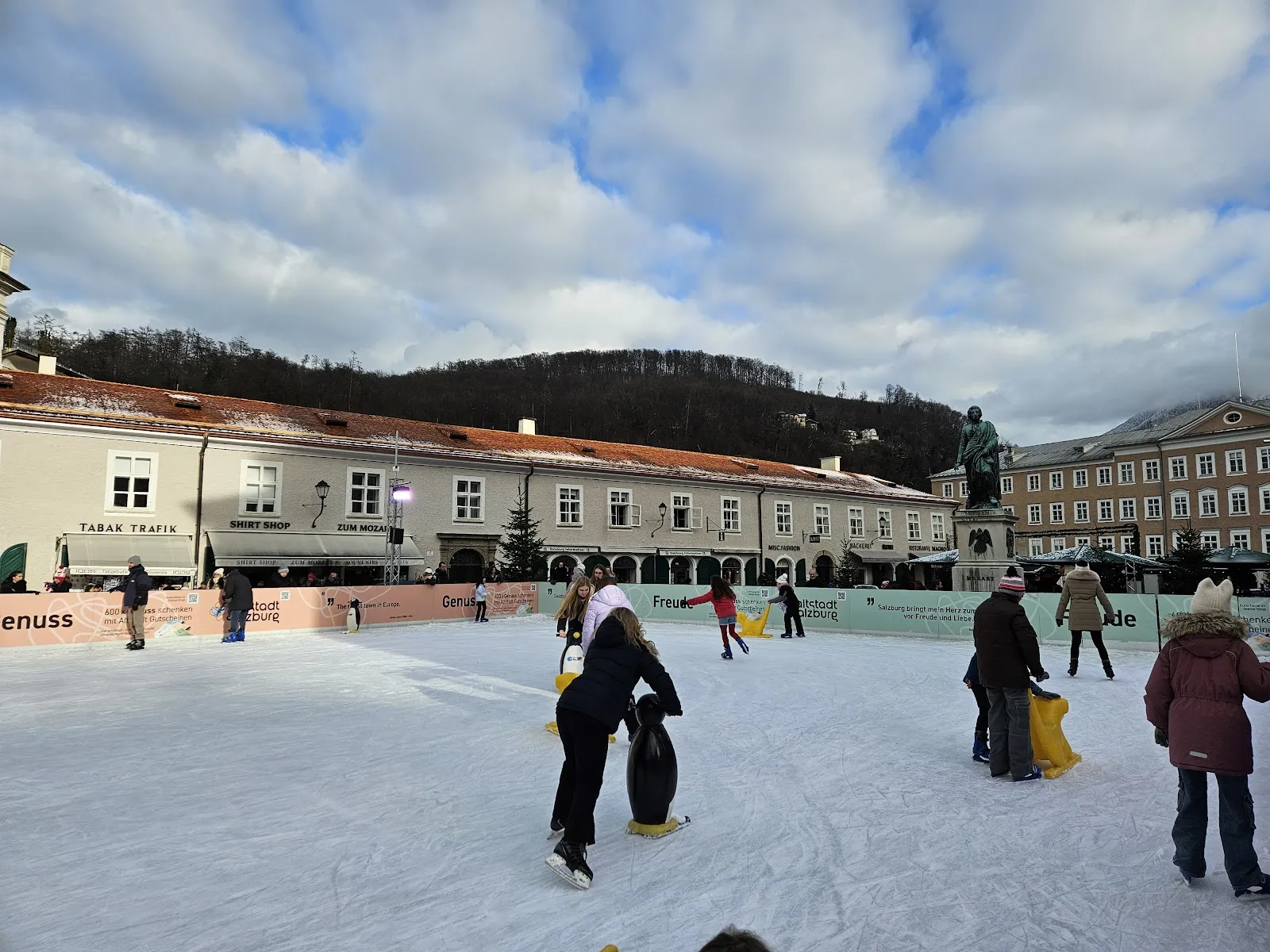 The Eiszauber rink on the Mozartplatz in Salzburg in daylight, with the Mozart monument and the old-town houses behind the skaters. — Eiszauber am Mozartplatz
