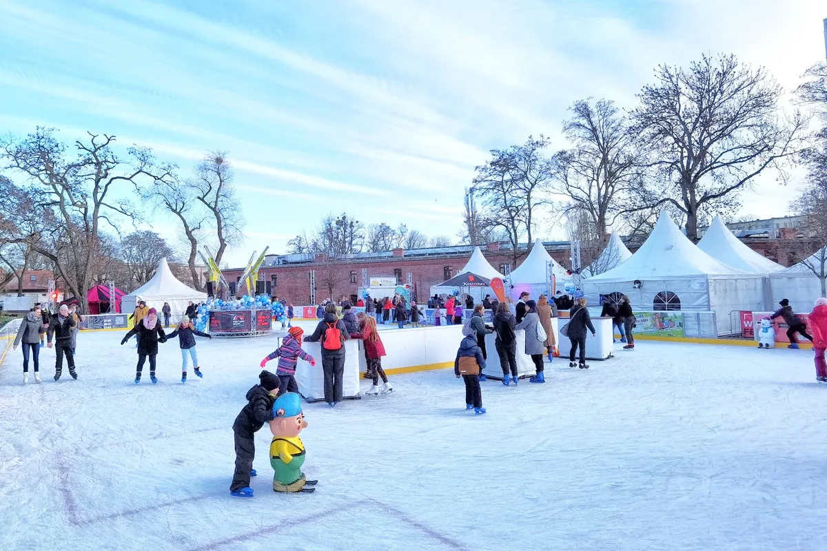 Schlittschuhläufer auf der Freiluft-Eisbahn der Eiszeit in der Festung Mark in Magdeburg an einem Wintertag, mit weißen Zelten und kahlen Bäumen rund um das Eis. – Eiszeit Festung Mark