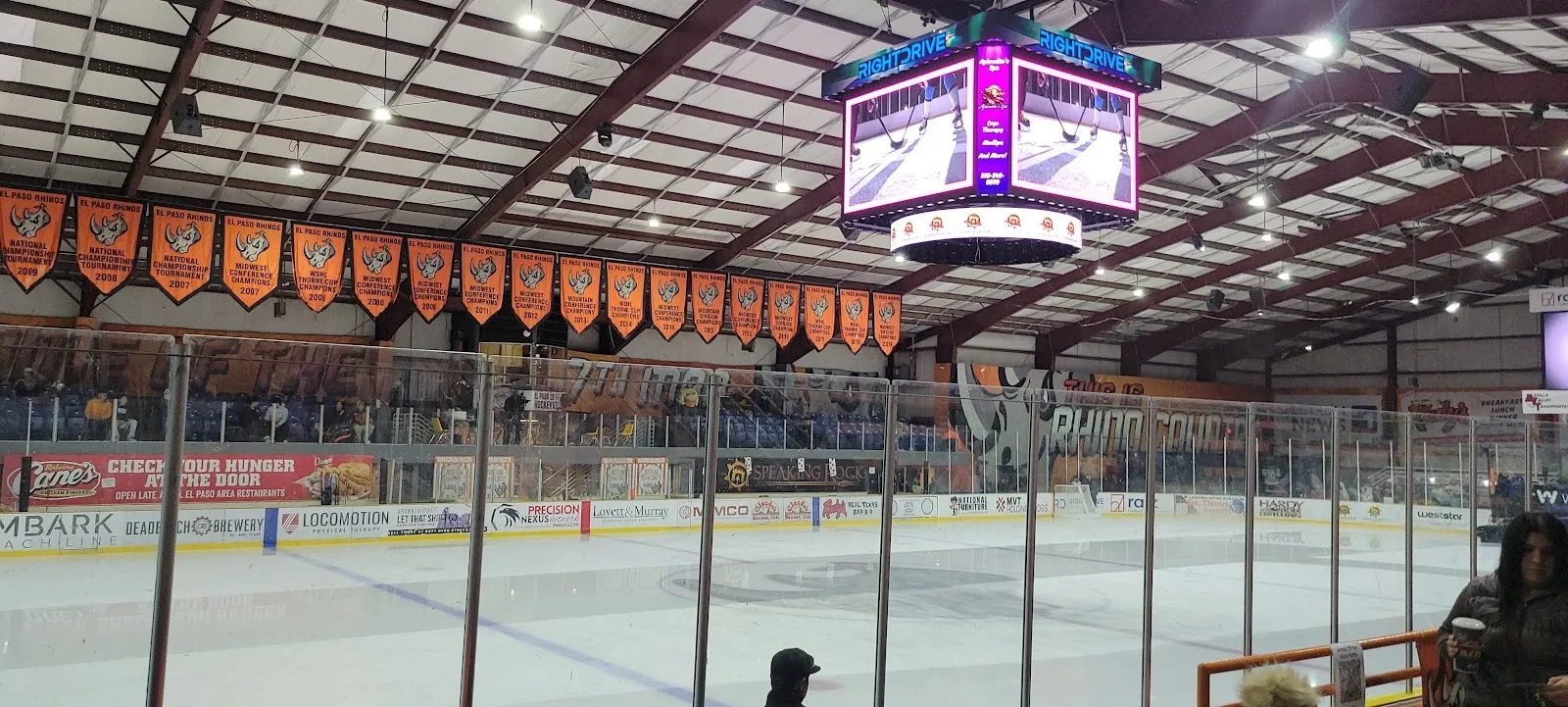 The coliseum ice seen from the seats, the sheet and rink glass under a scoreboard with orange banners hung from the roof. — El Paso County Events Center