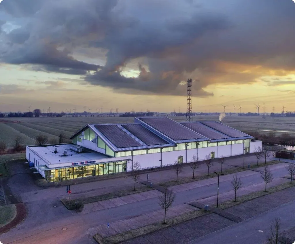 Aerial view of the Elbe Ice Stadion in Brokdorf at dusk, a long white hall with solar-panel roof in flat marshland — Elbe Ice Stadion