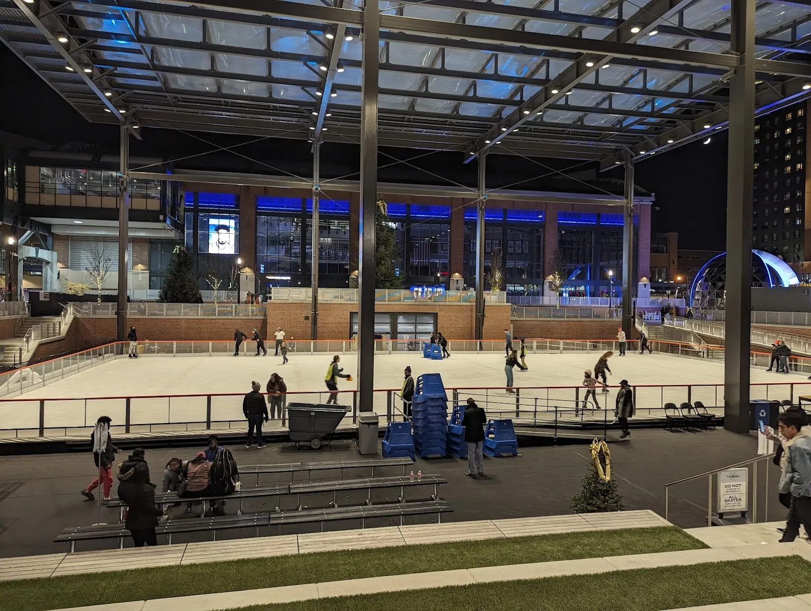The seasonal ice rink on Morris Bicentennial Plaza under a glass-and-steel canopy, with skaters spread across the ice, tiered benches in the foreground and a lit building on the far side. — Elevance Health Rink at Morris Bicentennial Plaza