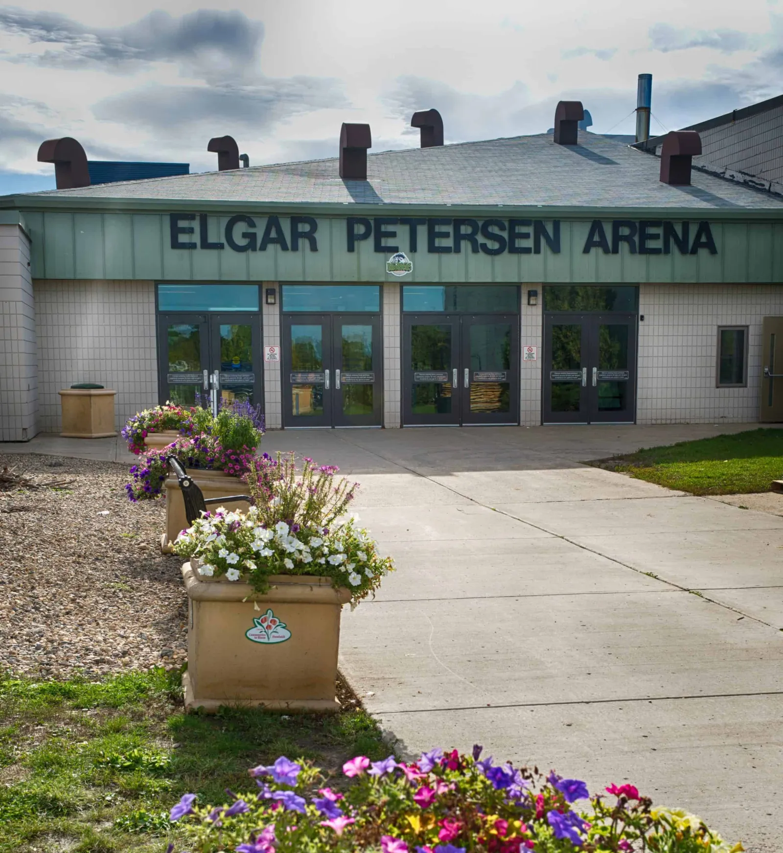 The Elgar Petersen Arena entrance with its name in large letters above four sets of glass doors, and flower planters along the walk. — Elgar Petersen Arena