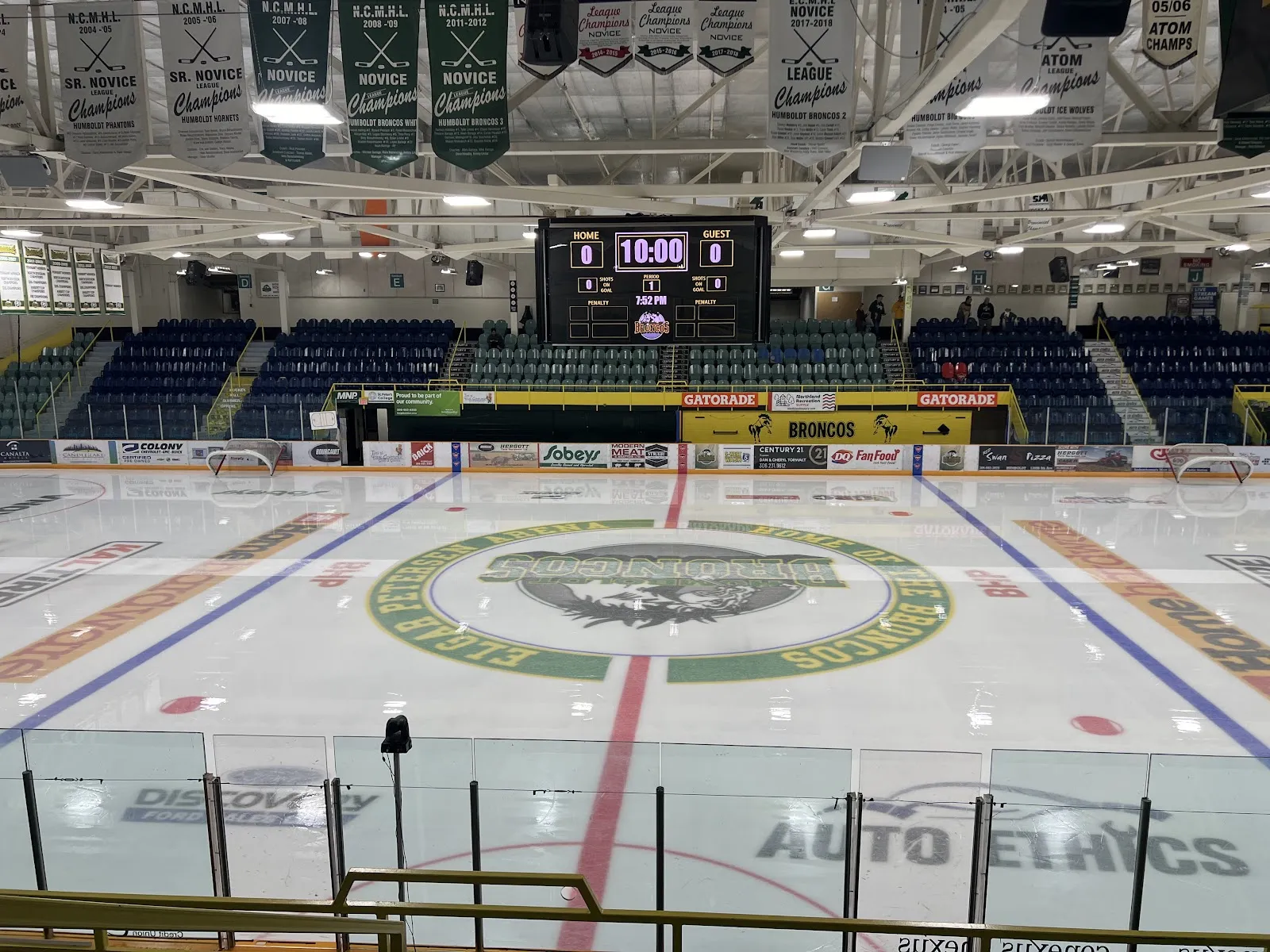 The empty Elgar Petersen Arena ice with the Humboldt Broncos logo at centre ice, blue seats, the scoreboard and championship banners overhead. — Elgar Petersen Arena